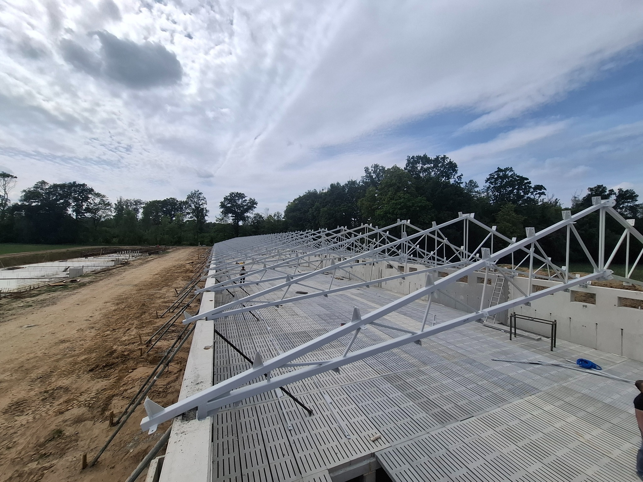 Konstrukcja hali w trakcie budowy: białe, metalowe elementy szkieletu na betonowym fundamencie, widok z góry, w tle drzewa i pochmurne niebo.