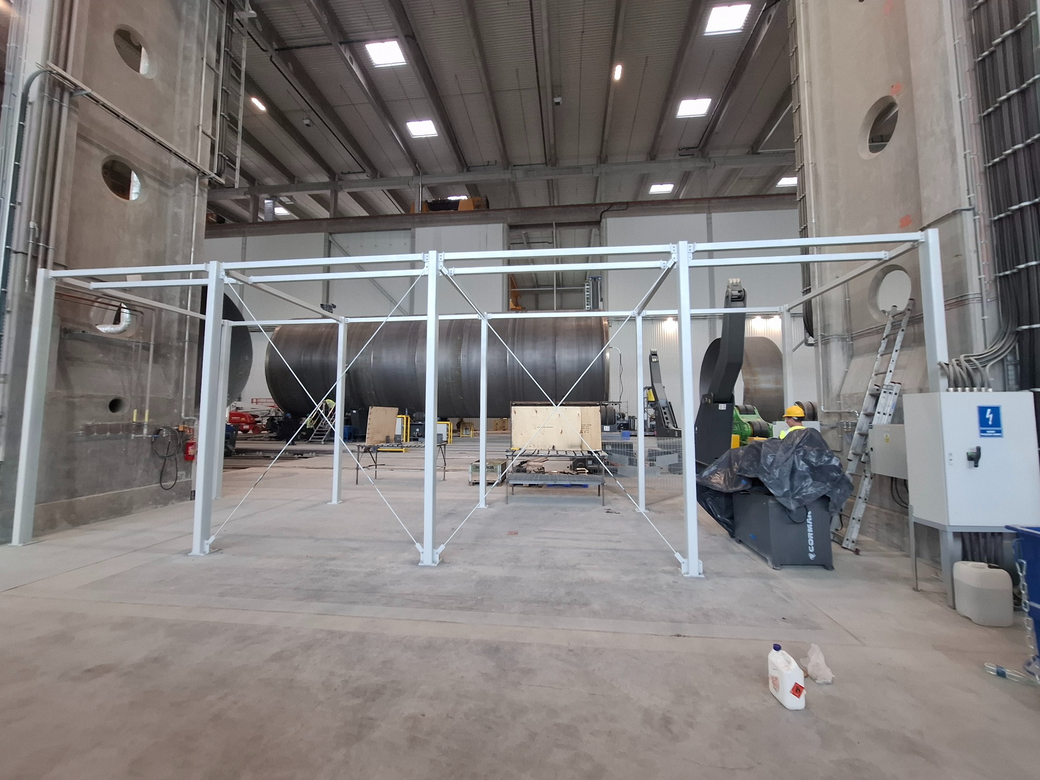 Wnętrze hali z konstrukcją stalową w trakcie montażu. Widoczne elementy mocujące, zbiornik i drabina. Oświetlenie górne, surowy beton na ścianach i podłodze.