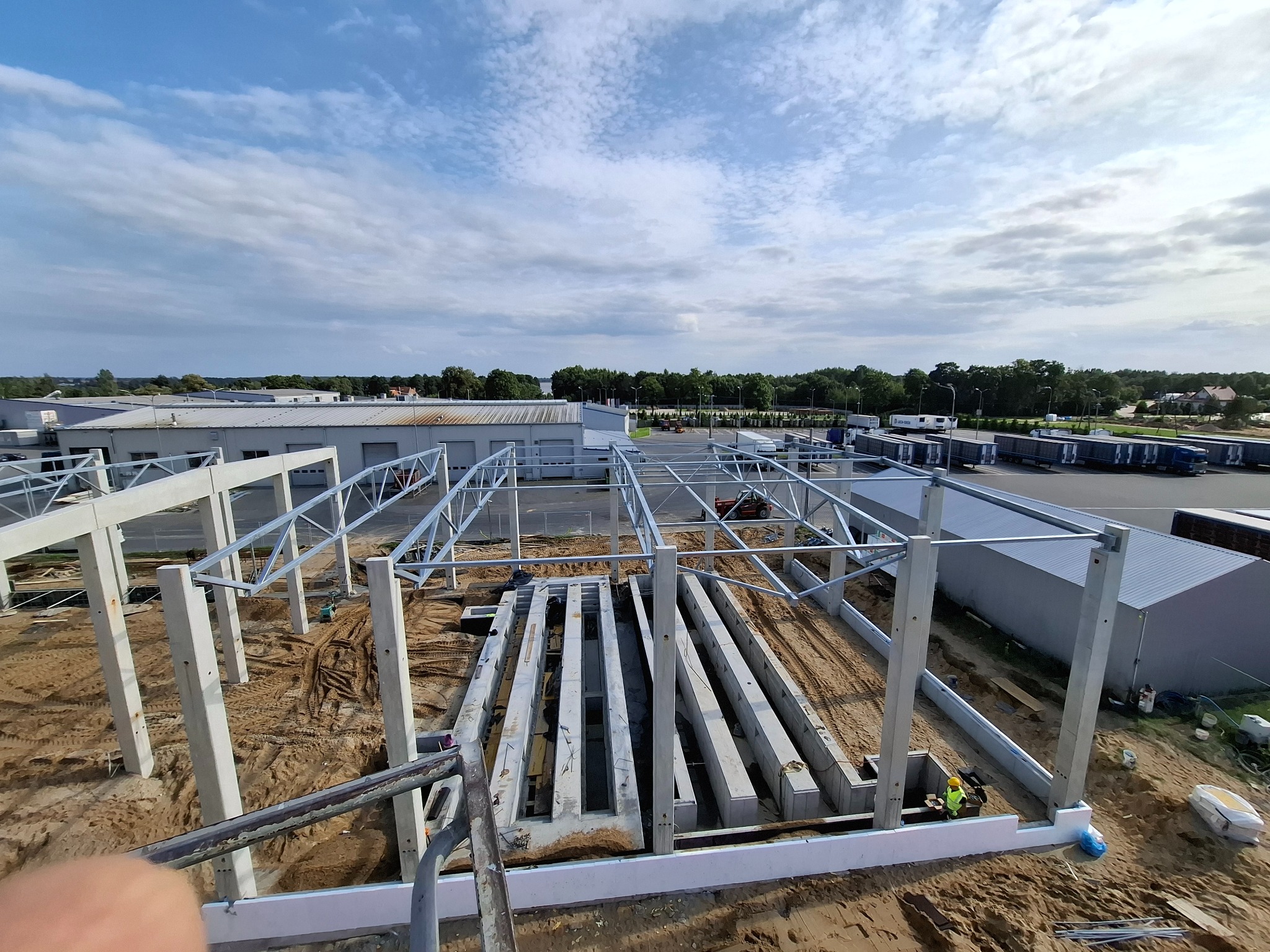 Konstrukcja hali w trakcie budowy: betonowe słupy i stalowa rama na tle błękitnego nieba z chmurami. Widoczne elementy zbrojenia i teren budowy.