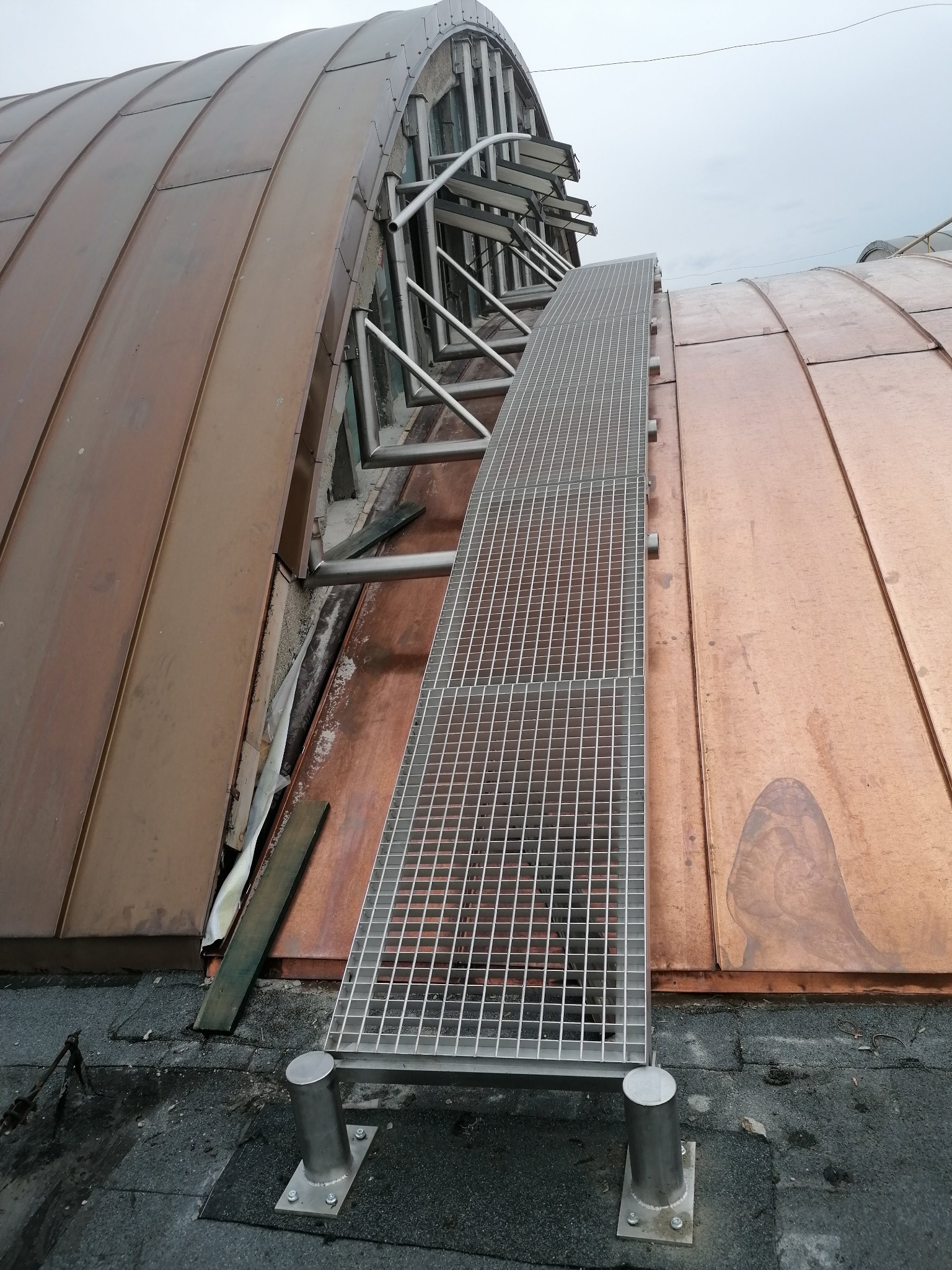 Metalowa kładka na miedzianym dachu łukowym, widok z dołu. Konstrukcja z kraty i rur, mocowana do dachu. Elementy montażowe widoczne. Nietypowe ujęcie.