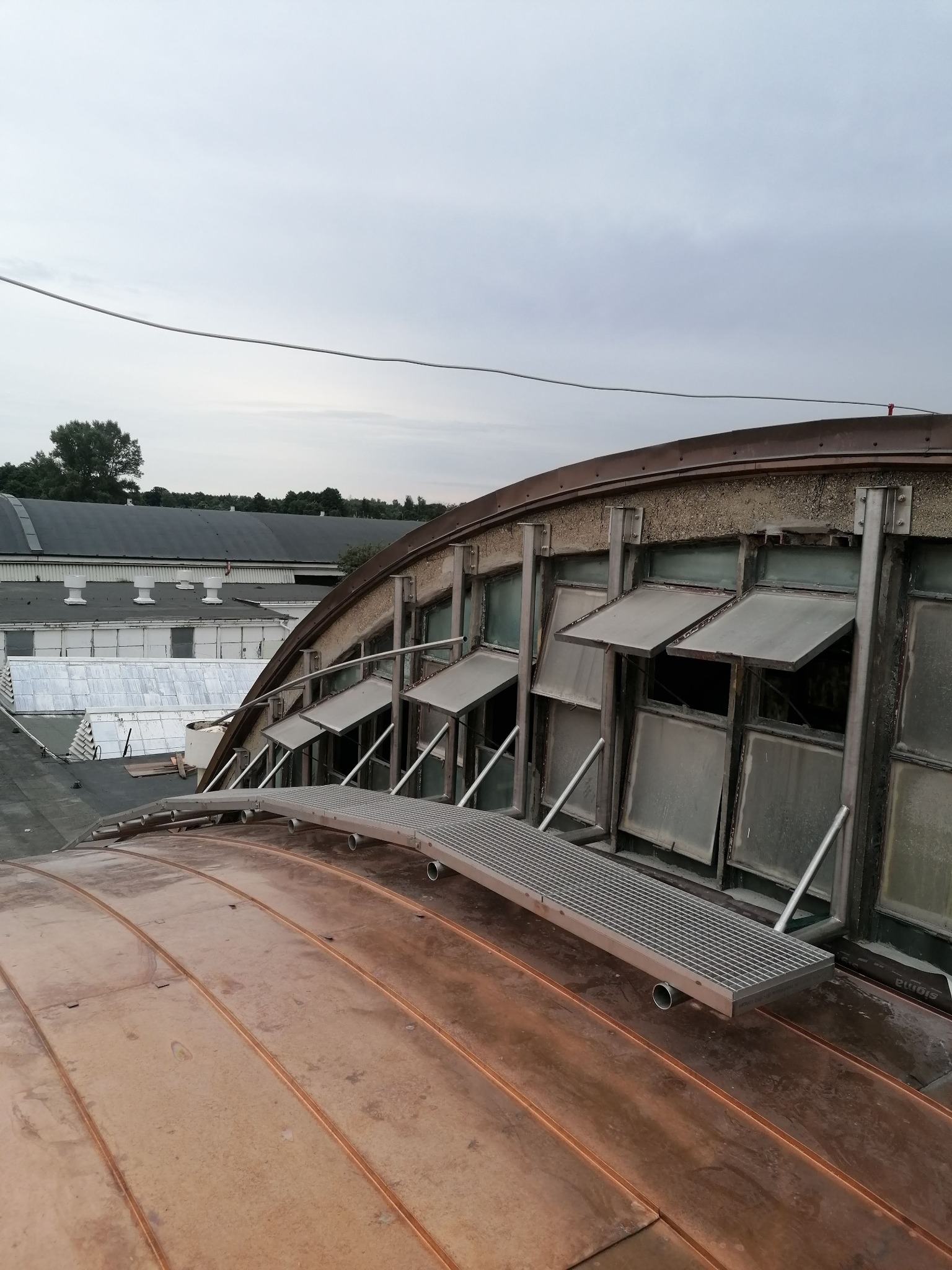 Miedziana kopuła z metalową kładką i oknami uchylnymi, widok z góry na tle zabudowań przemysłowych i pochmurnego nieba.