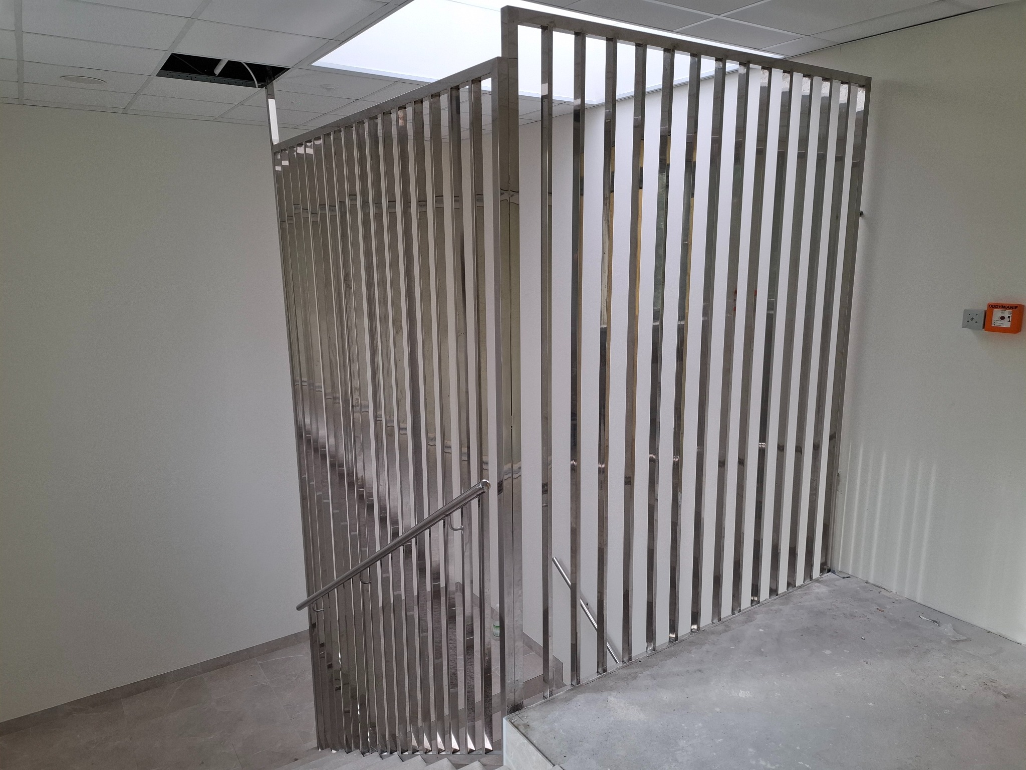 Nowoczesna, metalowa balustrada schodowa z pionowych profili, widok z góry na schody w jasnym wnętrzu. Minimalistyczny design, surowy beton na podłodze.