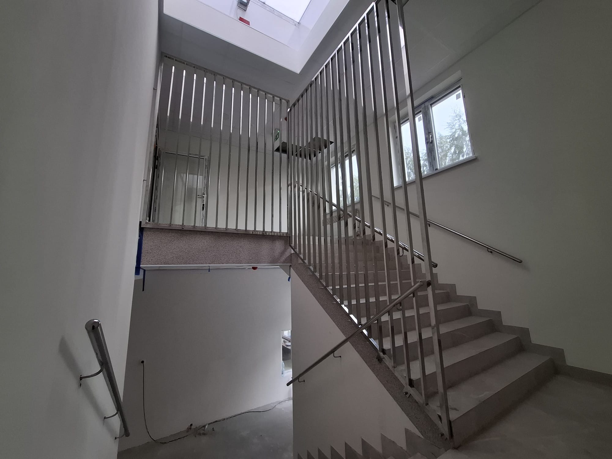 Wnętrze klatki schodowej z metalową balustradą o pionowych szczeblach, widok z dołu, oświetlenie naturalne z okna i świetlika dachowego.