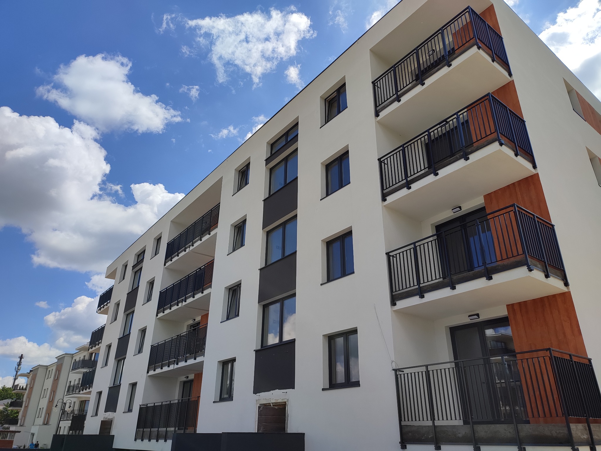 Nowoczesny budynek wielorodzinny z balkonami zabezpieczonymi czarnymi, metalowymi balustradami na tle błękitnego nieba z chmurami. Ujęcie z dołu, podkreślające wysokość budynku.