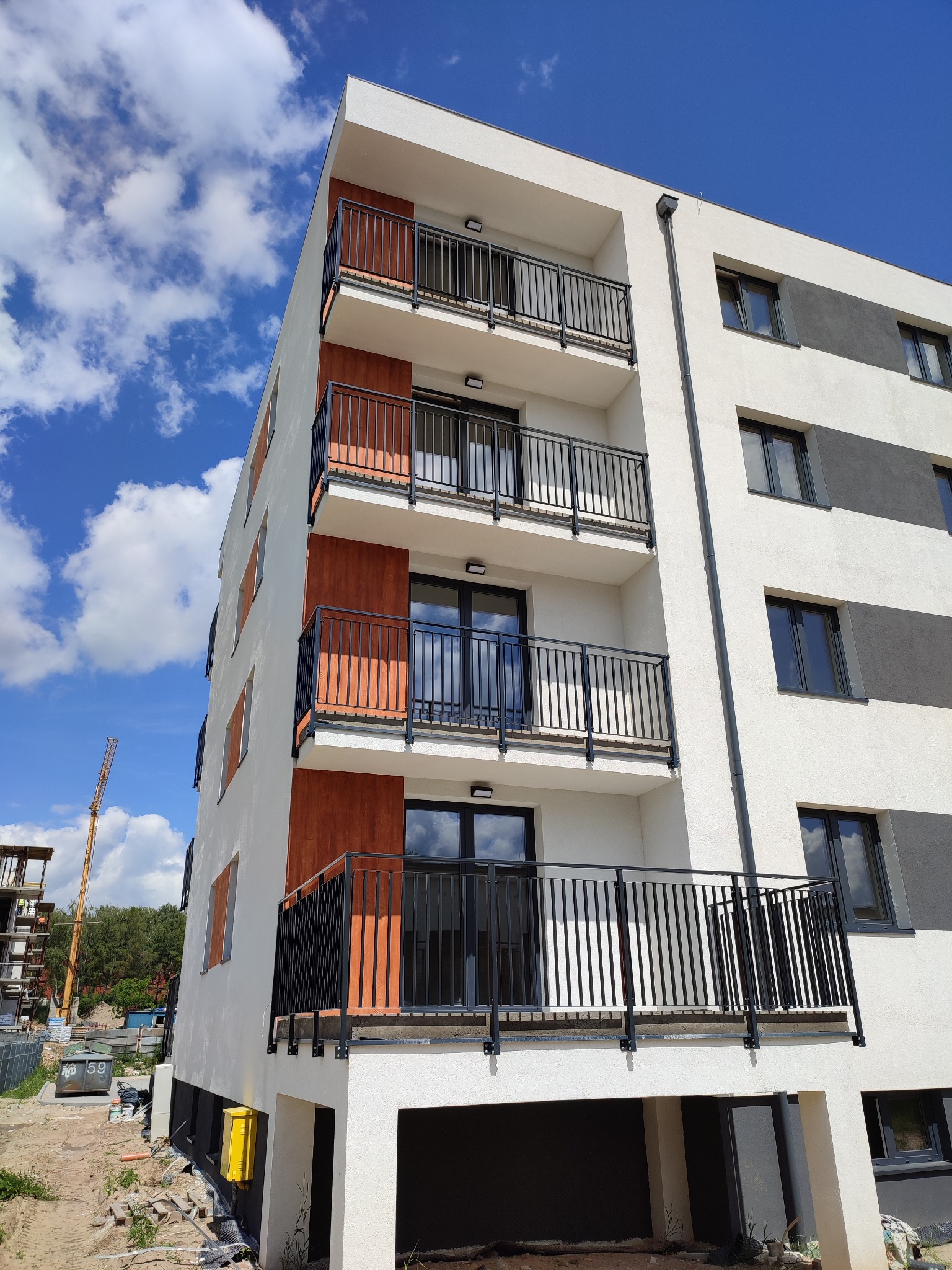 Nowoczesny budynek wielorodzinny z balkonami zabezpieczonymi czarnymi, metalowymi balustradami. Widok z dołu na elewację z elementami drewnopodobnymi i błękitne niebo.