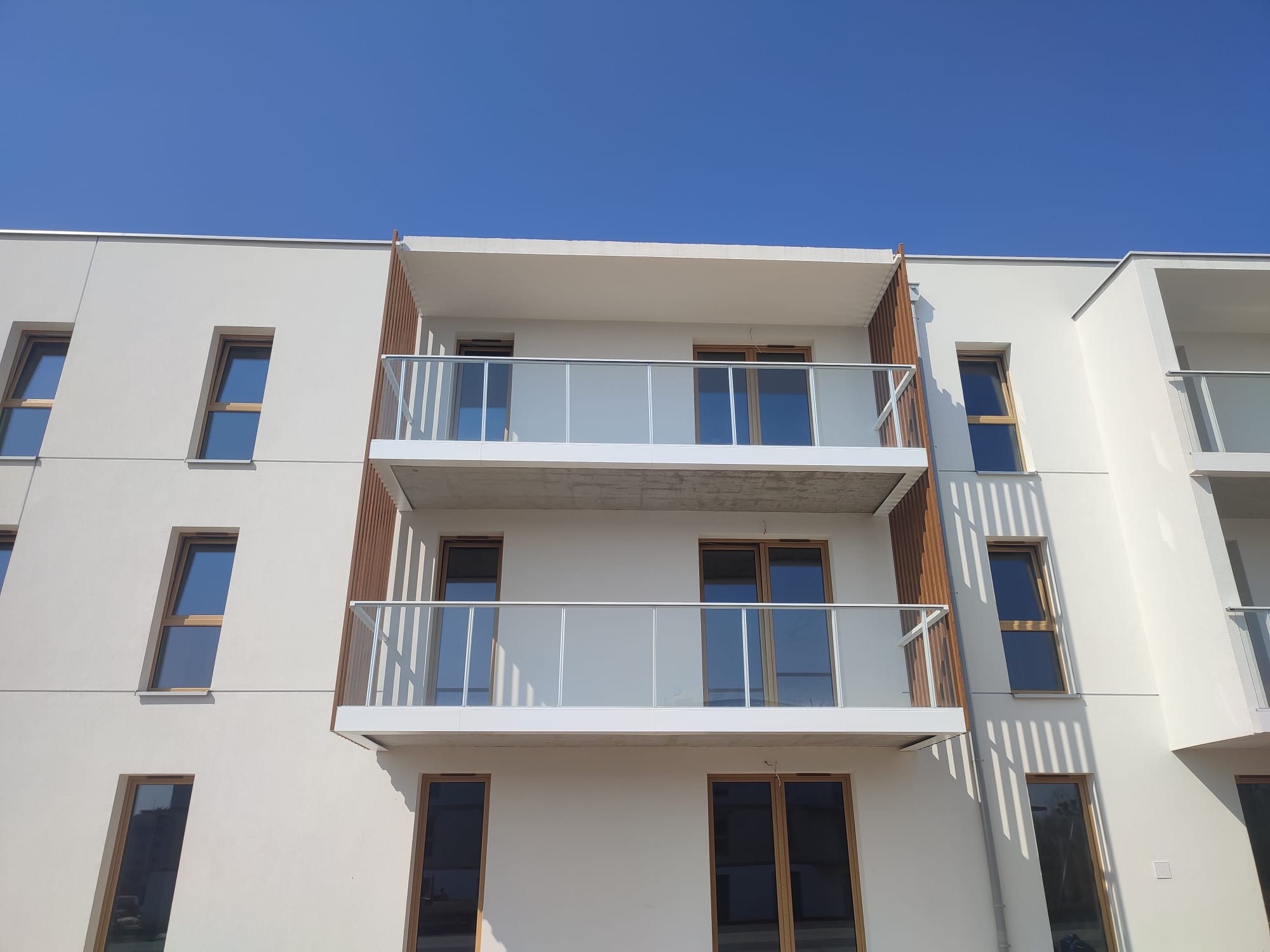 Nowoczesny budynek z białymi balkonami i szklanymi balustradami, drewniane elementy dekoracyjne, minimalistyczny design, błękitne niebo w tle.