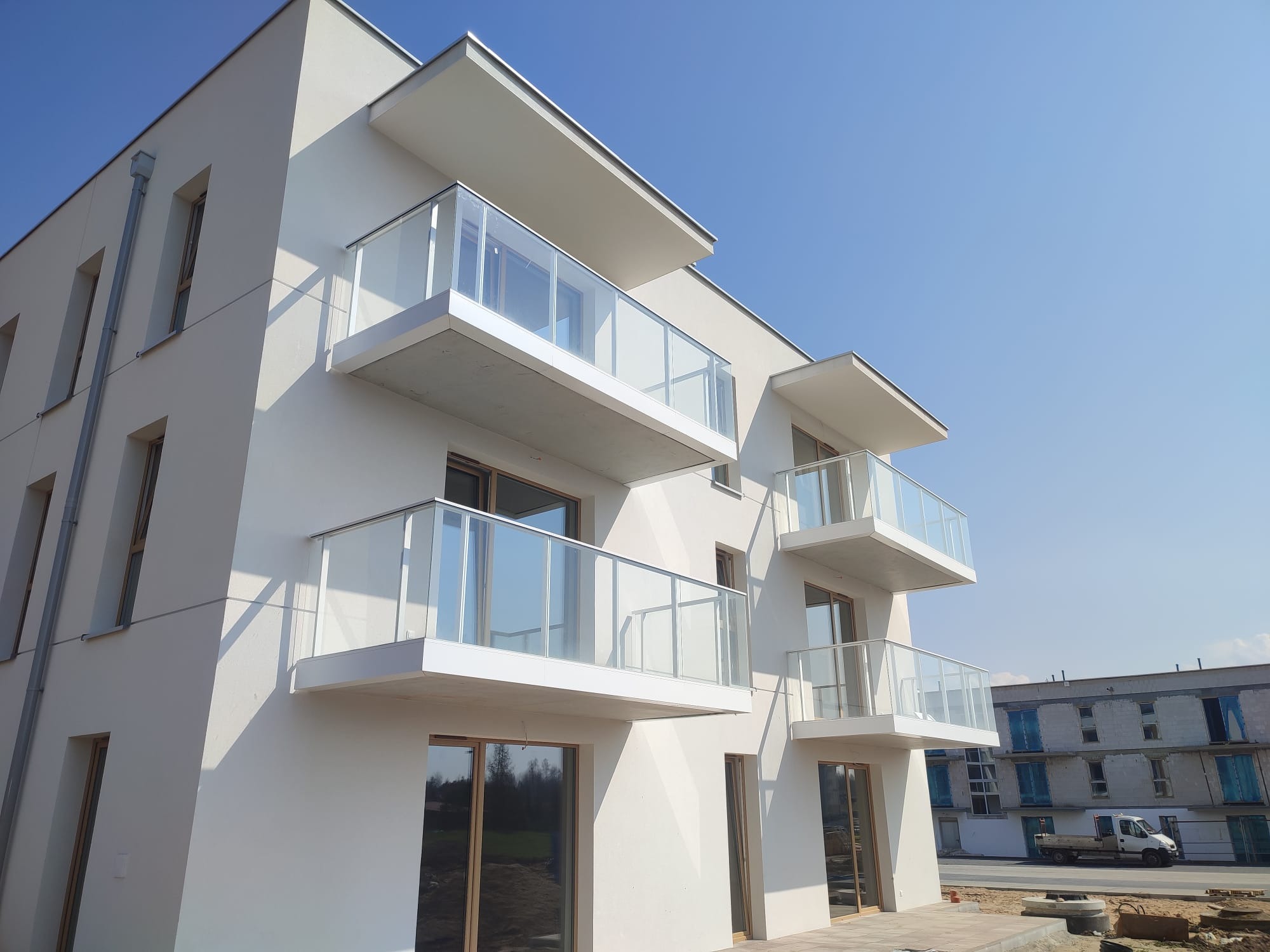 Nowoczesny budynek z balkonami z balustradami szklanymi, widok z dołu, jasne tło nieba. W tle budynek w trakcie budowy i samochód dostawczy.
