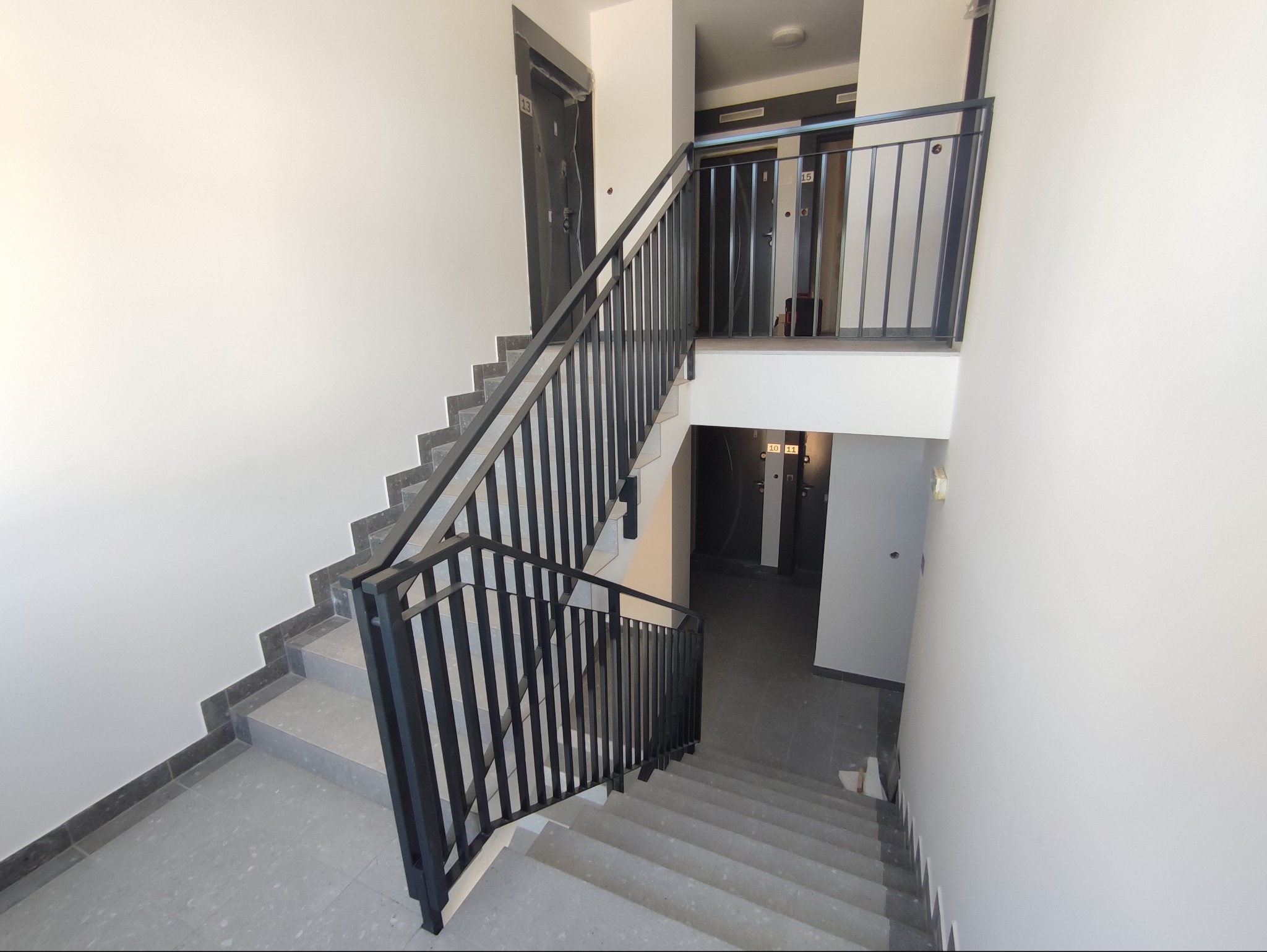Klatka schodowa w bloku z szarymi schodami i czarną, metalową balustradą. Widok z góry na schody prowadzące w dół. Drzwi do mieszkań z numerami na każdym piętrze.