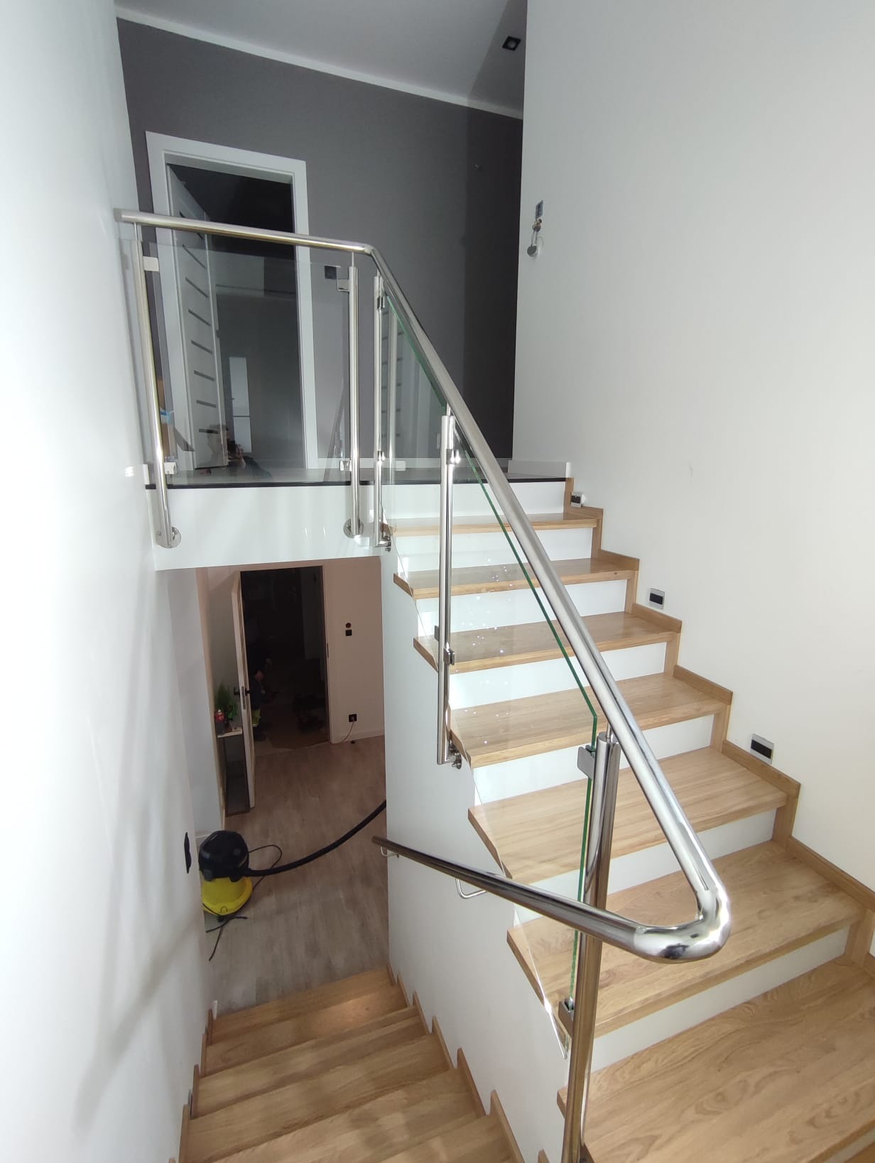 Nowoczesne schody z drewnianymi stopniami i balustradą ze stali nierdzewnej oraz szkła. Widok z góry na dół, z widocznym odkurzaczem na dole schodów.