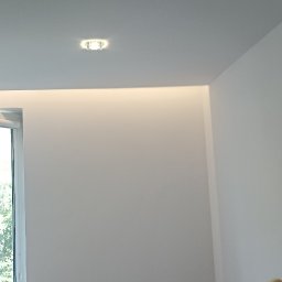 usługi budowlane - Wnętrze po malowaniu: białe ściany, sufit z oświetleniem punktowym i listwą LED, okno z grzejnikiem. W rogu widoczny fragment gąbki malarskiej i wiadro.