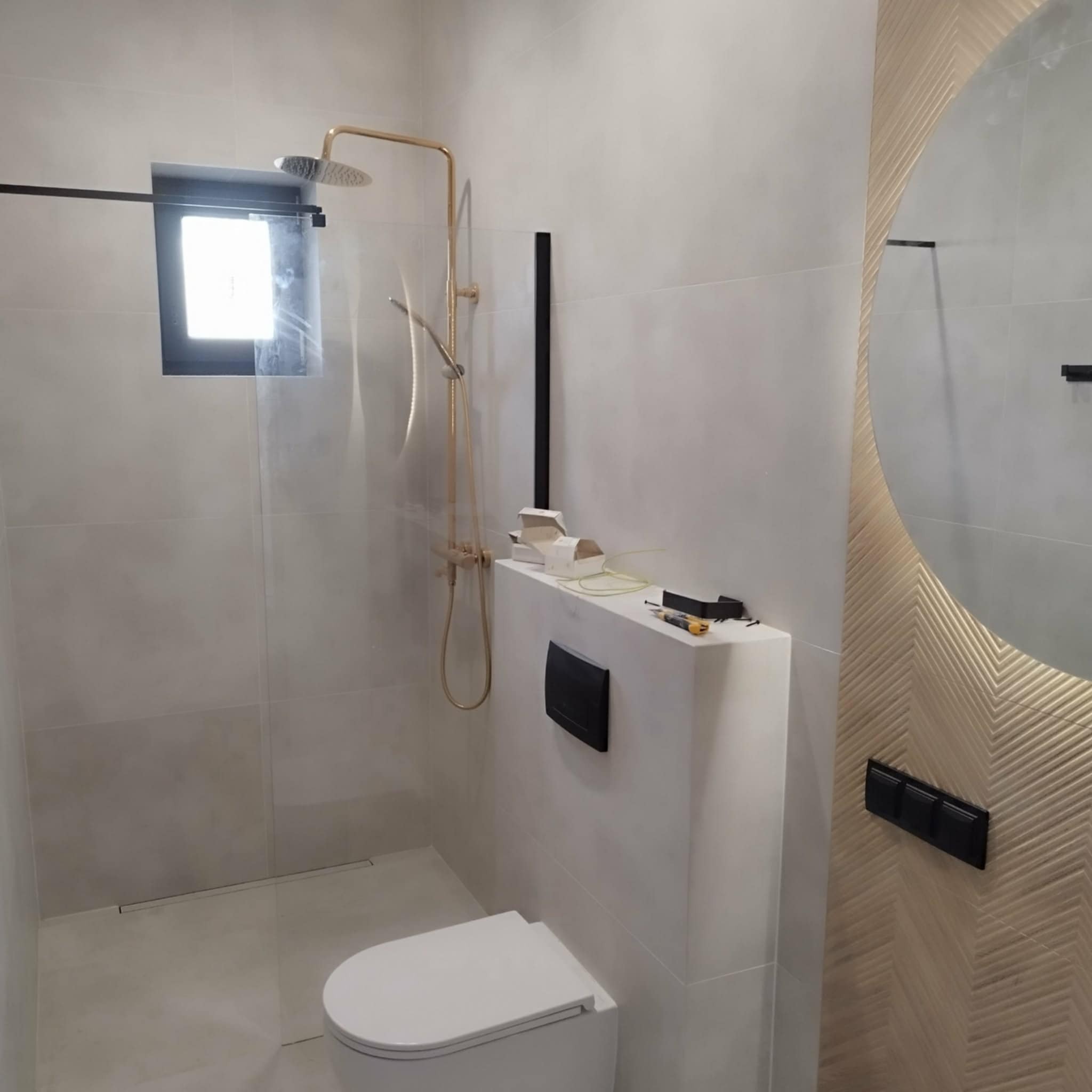 Nowoczesna łazienka w trakcie wykańczania: białe płytki, złota armatura, okrągłe lustro z dekoracyjną ramą. Widoczne narzędzia i akcesoria montażowe.