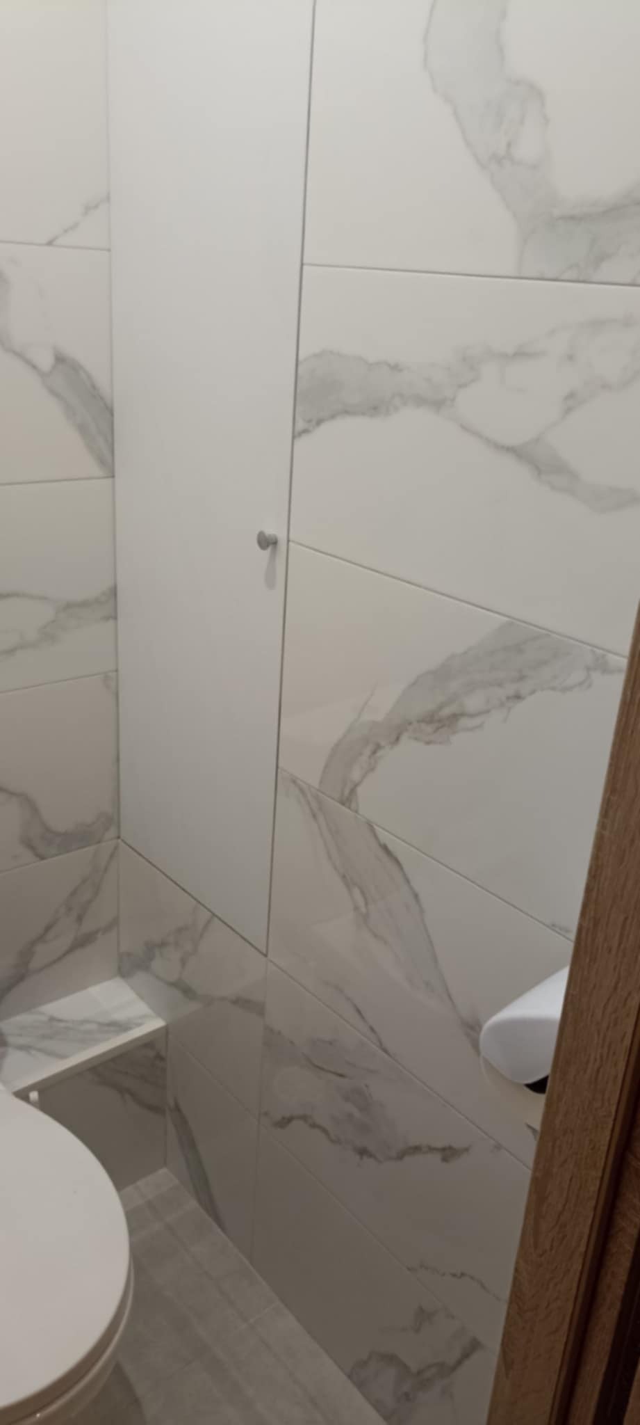 Wykończona łazienka z białymi, marmurkowymi płytkami, ukryte drzwi rewizyjne z małym, okrągłym uchwytem i fragment toalety w dolnym rogu kadru.