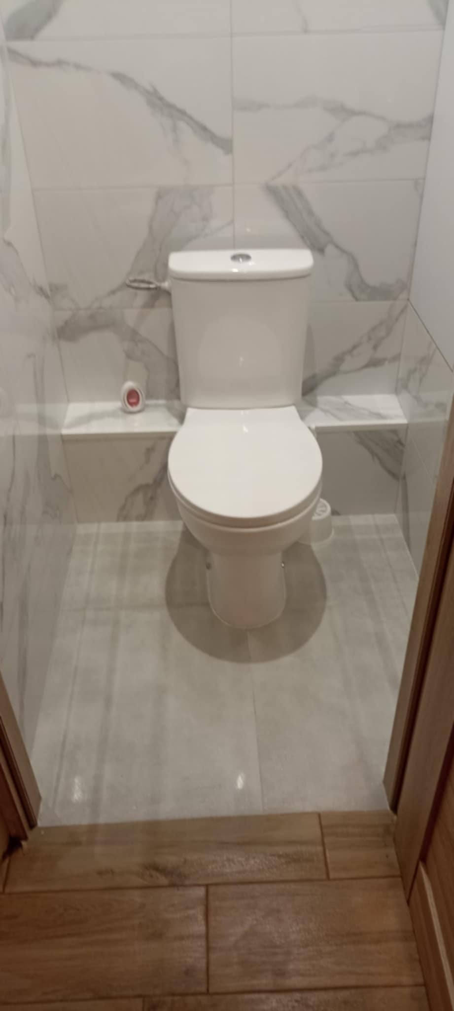 Biała toaleta w wykończonej łazience z marmurowymi płytkami na ścianach i podłodze. Widoczne drewniane wykończenie drzwi.