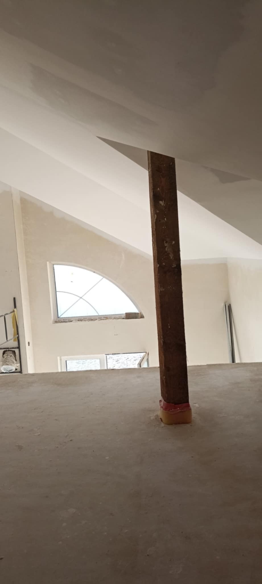 Wnętrze w trakcie prac wykończeniowych: surowe ściany, drewniany słup zabezpieczony folią, okno o nietypowym kształcie, widoczne elementy rusztowania.