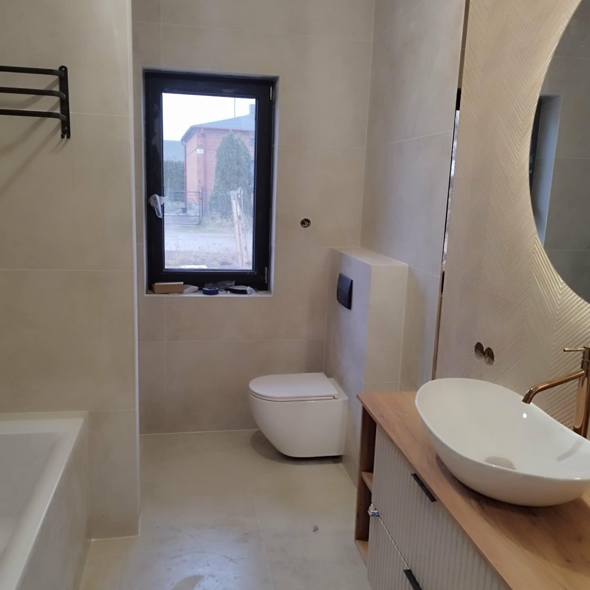 Nowoczesna łazienka w beżowych odcieniach z wiszącą toaletą, umywalką na drewnianym blacie i dużym lustrem z podświetleniem. Widoczne płytki na ścianach i podłodze.