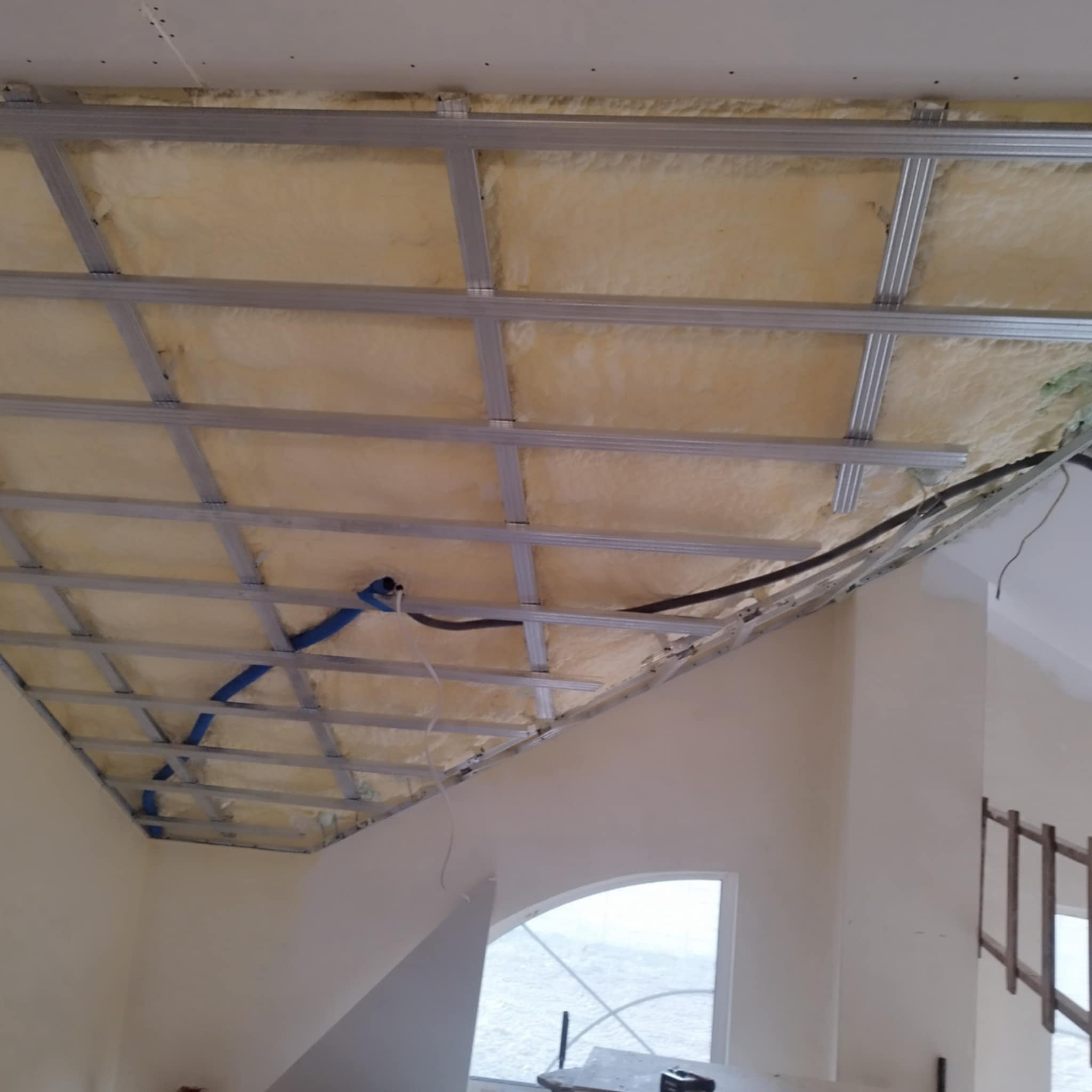 Sufit podwieszany z izolacją z wełny mineralnej i metalową konstrukcją, widok z dołu, w tle okno i fragment ściany. Etap prac wykończeniowych.