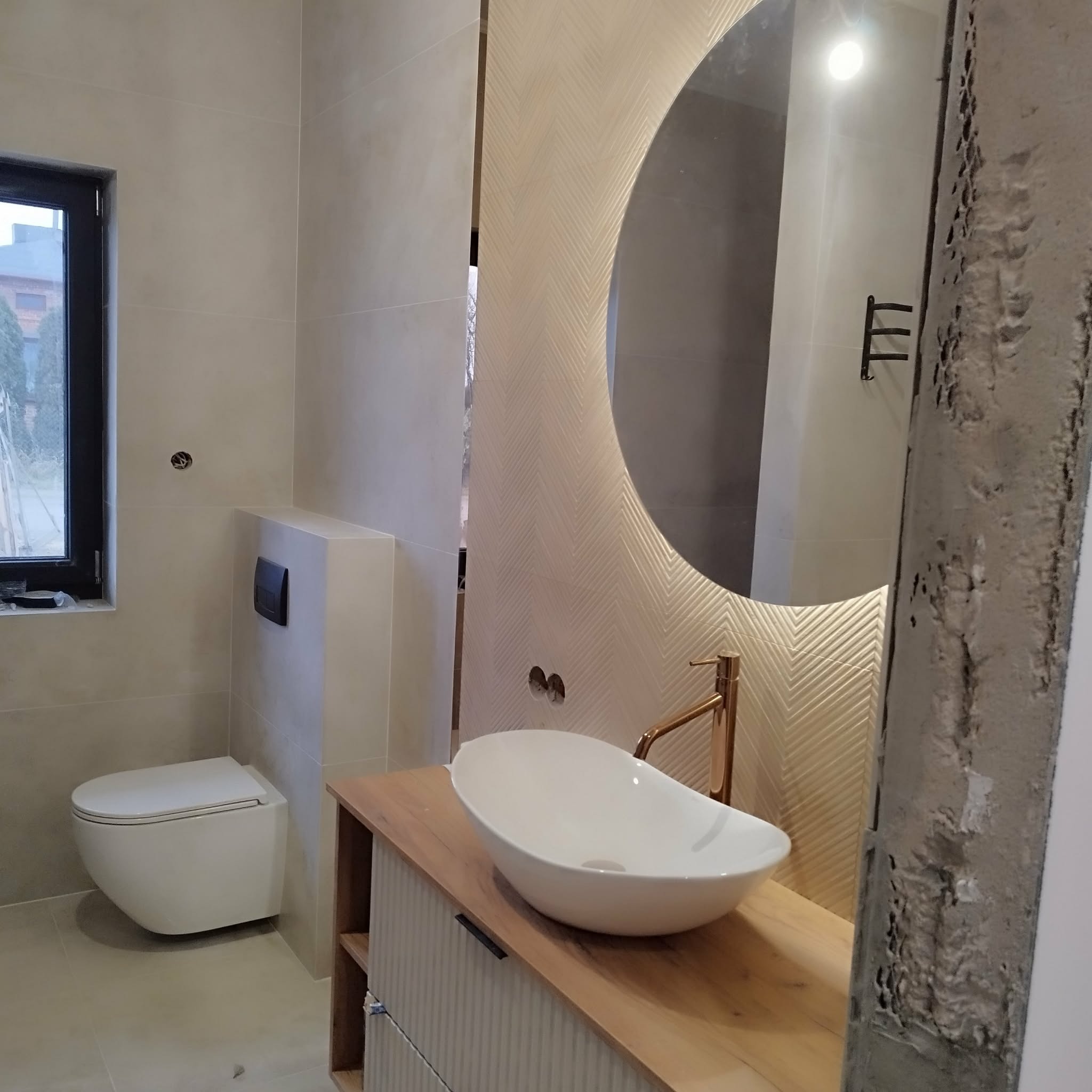 Nowoczesna łazienka z owalnym lustrem z podświetleniem LED, białą umywalką nablatową i złotą armaturą. Beżowe płytki i drewniane elementy wykończenia.