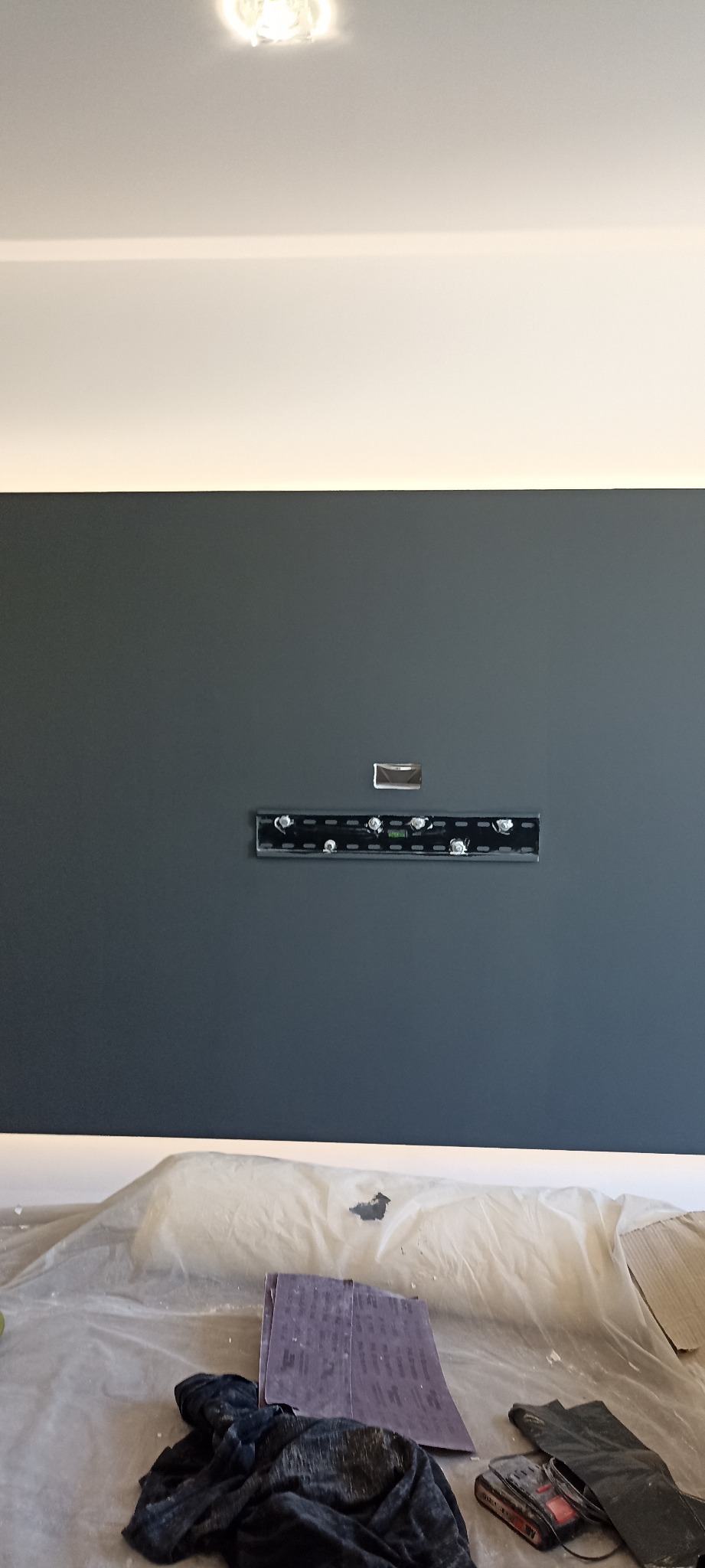 Ściana pomalowana na szaro z zamontowaną listwą do telewizora. Na podłodze folia ochronna, papier ścierny i narzędzia malarskie.