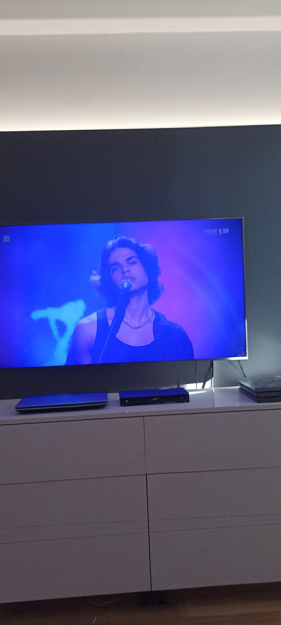 Telewizor LCD na szafce RTV, wyświetlający koncert w niebieskiej tonacji. Ściana pomalowana na szaro, biały sufit. Widoczne urządzenia elektroniczne.