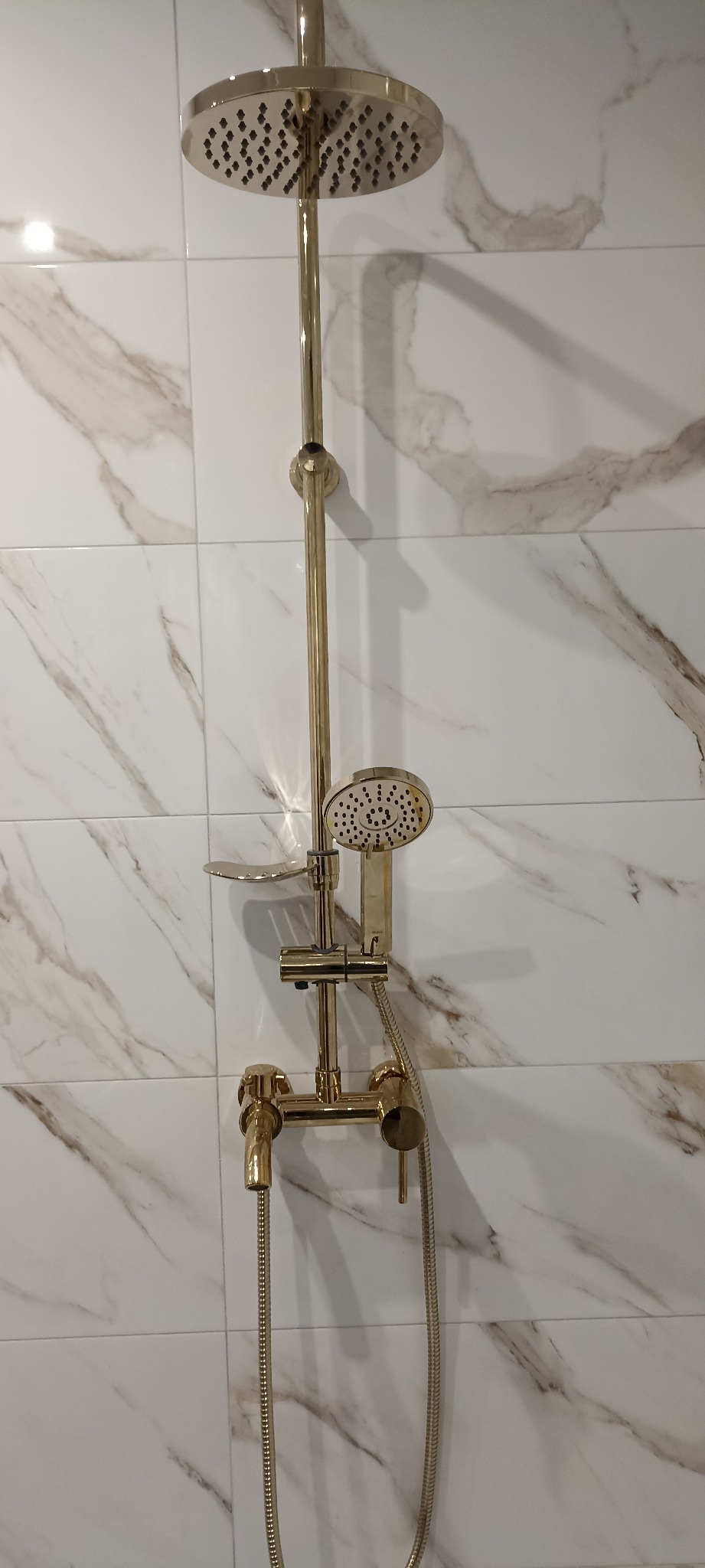 Złota bateria prysznicowa z deszczownicą na tle białych płytek imitujących marmur. Nowoczesny design łazienki po remoncie.