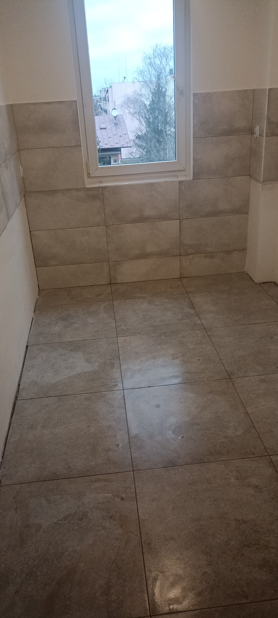 Wykończona łazienka z beżowymi płytkami na ścianach i podłodze, widok na okno z widokiem na budynki i drzewa. Minimalistyczny design.