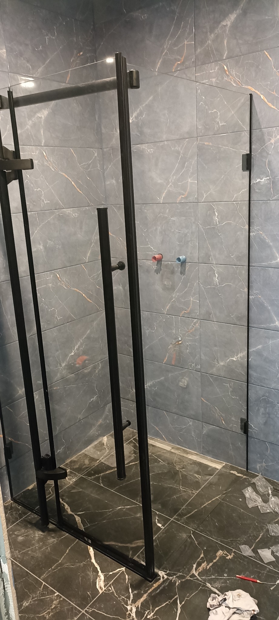 Nowoczesna kabina prysznicowa z czarnymi profilami na tle płytek imitujących marmur. Widoczne elementy instalacji wodnej.