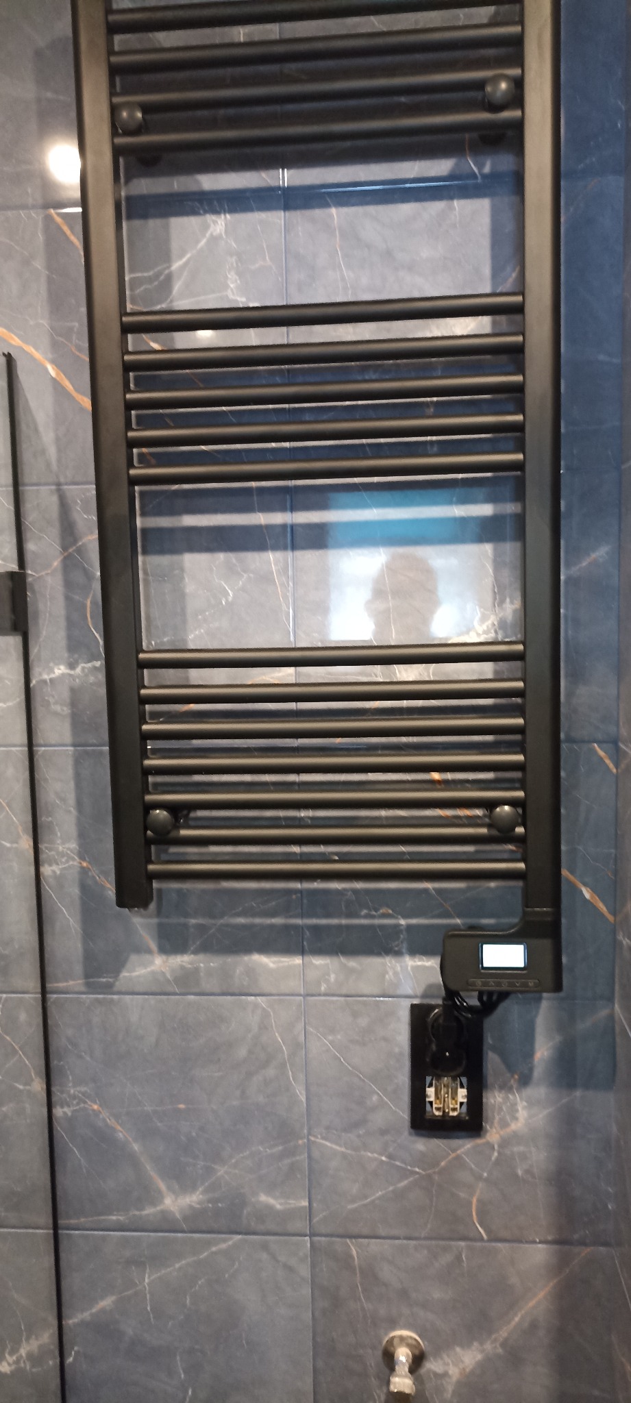 Czarny, drabinkowy grzejnik łazienkowy z elektronicznym sterownikiem, zamontowany na ścianie wyłożonej szarymi płytkami imitującymi marmur z delikatnymi, złotymi żyłkami.