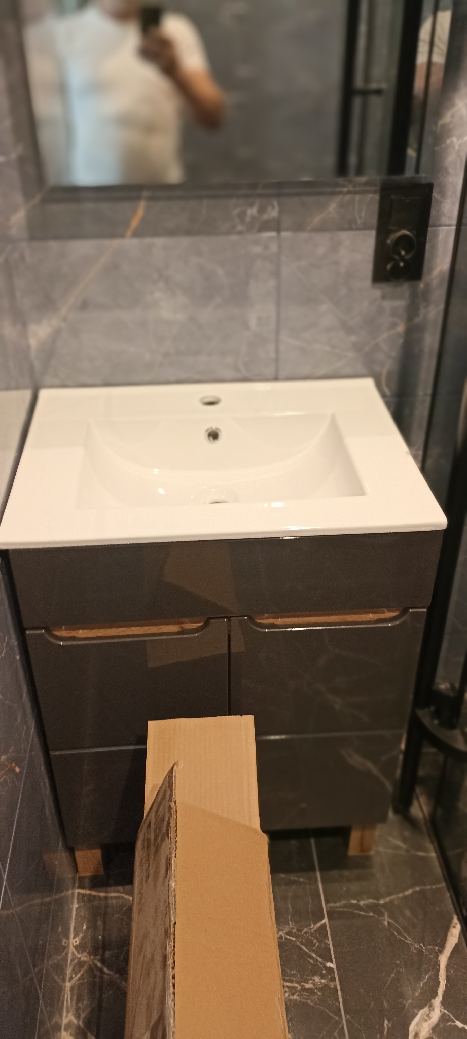 Nowoczesna umywalka z szafką w łazience wykończonej płytkami imitującymi marmur. Widoczny fragment lustra i kartonowe opakowanie na podłodze.