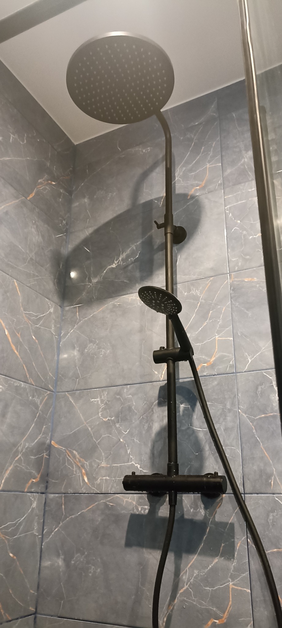 Nowoczesny, czarny zestaw prysznicowy z deszczownicą i słuchawką, zamontowany na ścianie z szarych płytek imitujących marmur w wykończonej łazience.