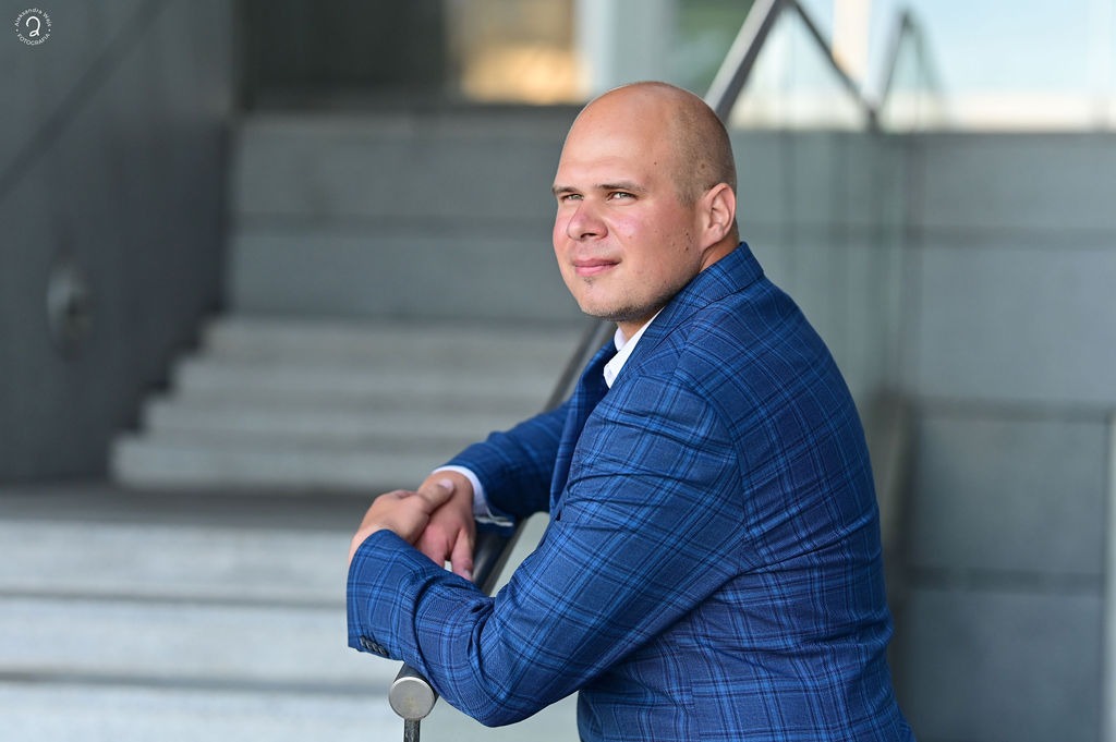 Portret mężczyzny w niebieskiej marynarce w kratę, opierającego się o metalową barierkę na tle schodów i szklanej fasady budynku.