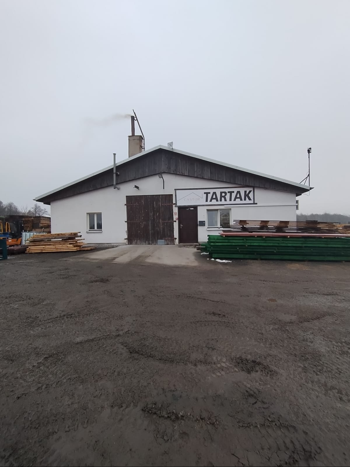 Budynek tartaku z widocznym stosem tarcicy. Na budynku szyld z nazwą firmy. Z komina unosi się dym. Pochmurny dzień.