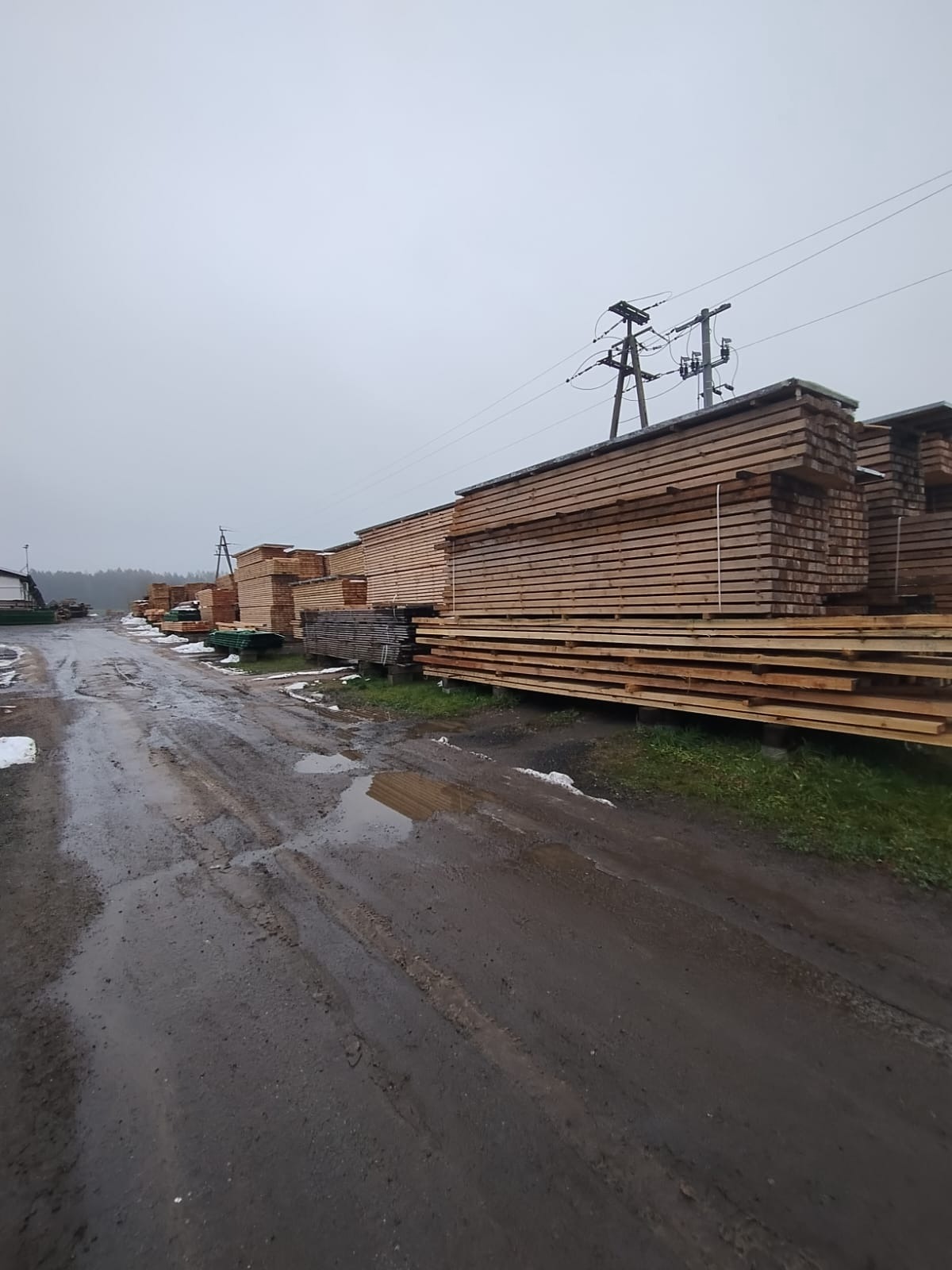 Stosy tarcicy na placu pod pochmurnym niebem, widoczne ślady opon na błotnistej drodze, w tle las i słupy energetyczne.