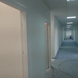 T.P Building Services - Pomalowany na biało korytarz biurowy z niebieską podłogą, widok w głąb z otwartymi drzwiami po obu stronach. Widoczne ślady po remoncie.