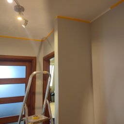 T.P Building Services - Przygotowanie do odświeżenia ścian: szara ściana zabezpieczona żółtą taśmą malarską, drabina aluminiowa i oświetlenie sufitowe. Widoczne drzwi z szybami.