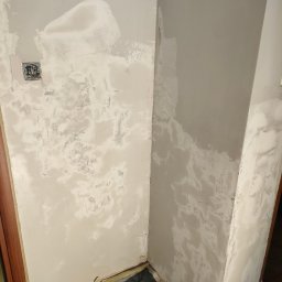 T.P Building Services - Narożnik pokoju w trakcie szpachlowania. Widoczne ślady po nałożonej gładzi gipsowej, przygotowanie do malowania. Zabezpieczona folią podłoga i listwy przypodłogowe.