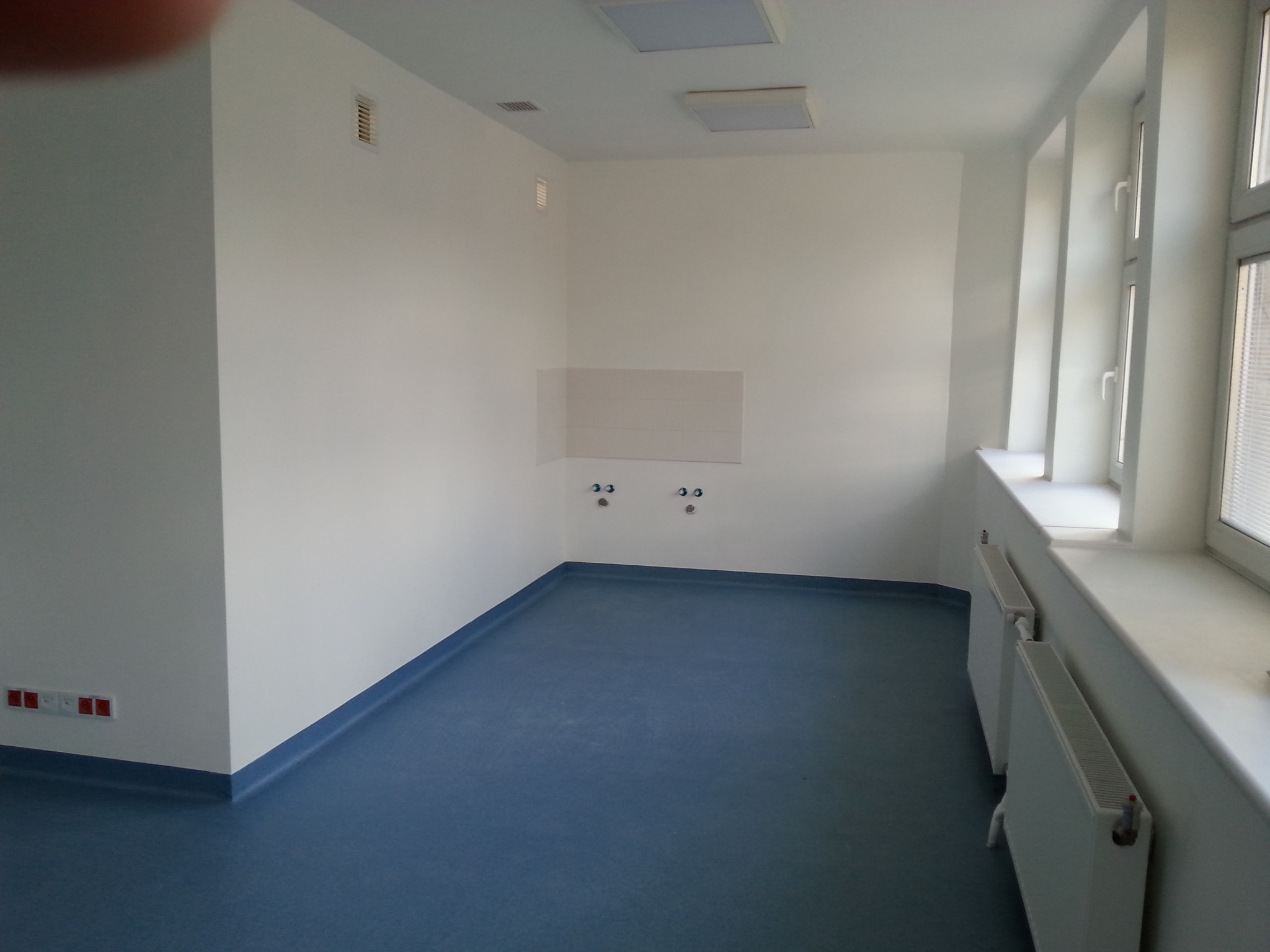 Wnętrze po remoncie: białe ściany, niebieska podłoga, grzejniki pod oknem. Widoczne gniazdka i instalacja wodna. Minimalistyczny styl, jasne oświetlenie.
