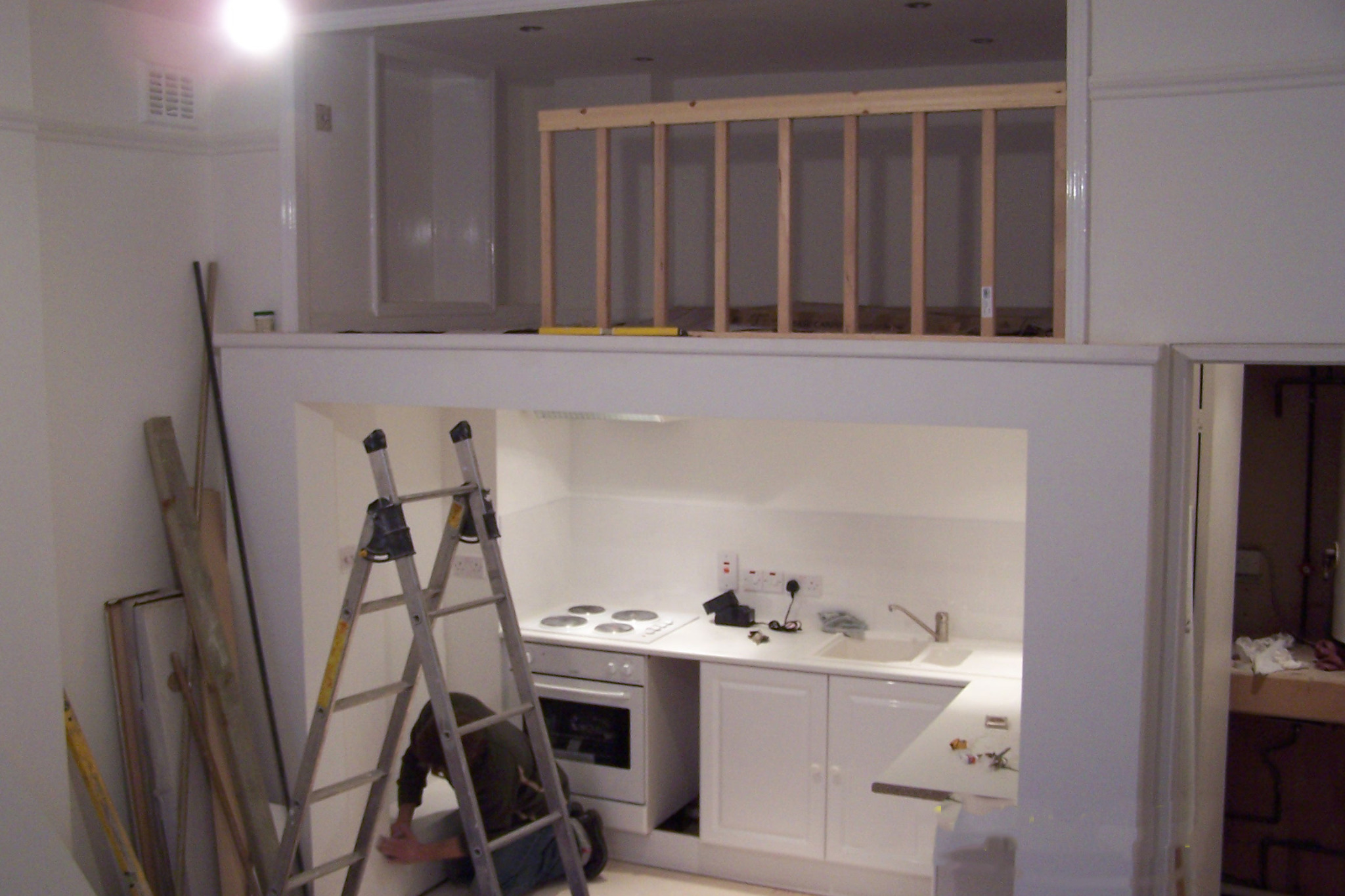 Remont kuchni: mężczyzna na drabinie maluje dolną część ściany. Widoczne materiały budowlane, białe meble kuchenne i antresola z drewnianą barierką.