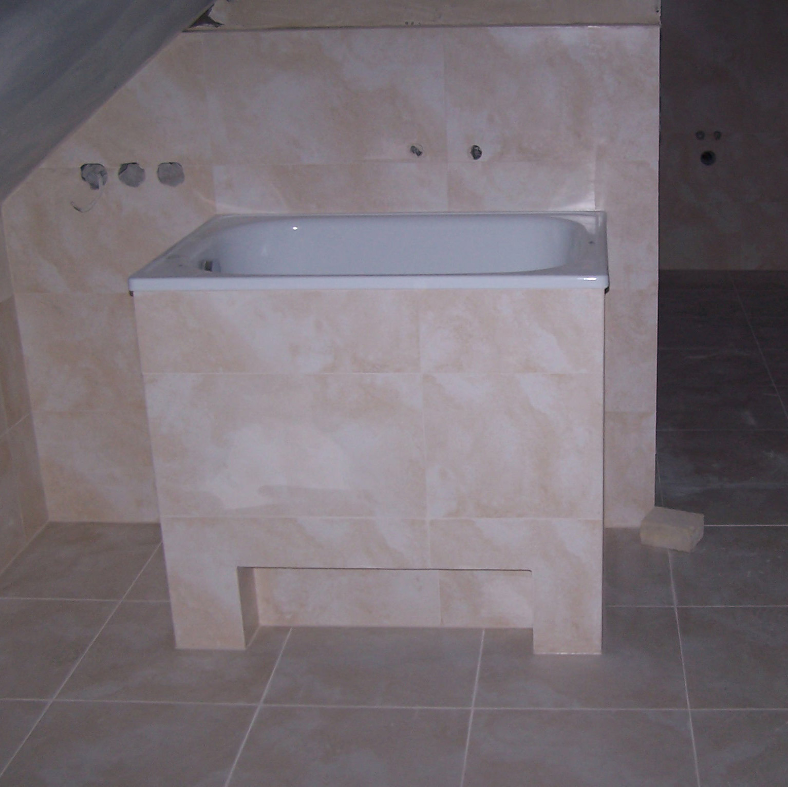 Wykończona obudowa wanny beżowymi płytkami w łazience na poddaszu. Widoczne otwory na baterię i fragment podłogi z płytek. Neutralna kolorystyka.