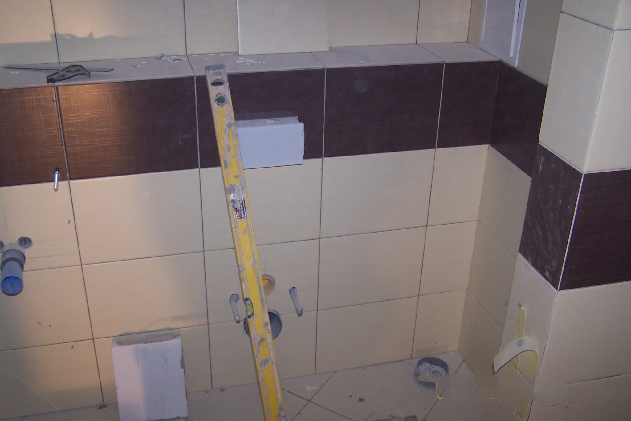 Wnętrze łazienki w trakcie układania płytek, widoczna poziomica oparta o ścianę, fragmenty płytek i instalacji. Prace glazurnicze w toku.