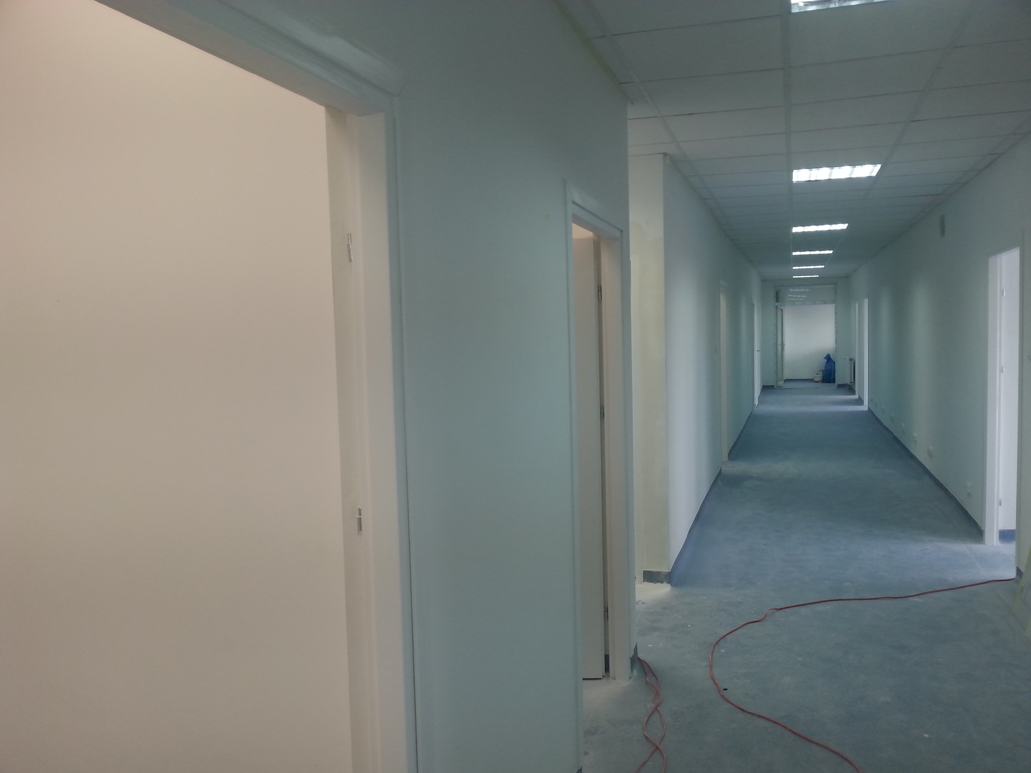 Pomalowany na biało korytarz biurowy z niebieską podłogą, widok w głąb z otwartymi drzwiami po obu stronach. Widoczne ślady po remoncie.