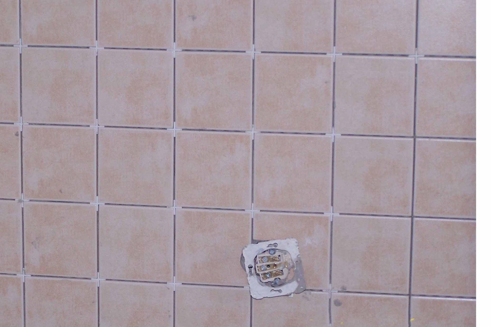 Ściana z beżowych, kwadratowych płytek z krzyżykami dystansowymi. W jednym miejscu widoczne gniazdko elektryczne bez osłony, w trakcie montażu płytek.