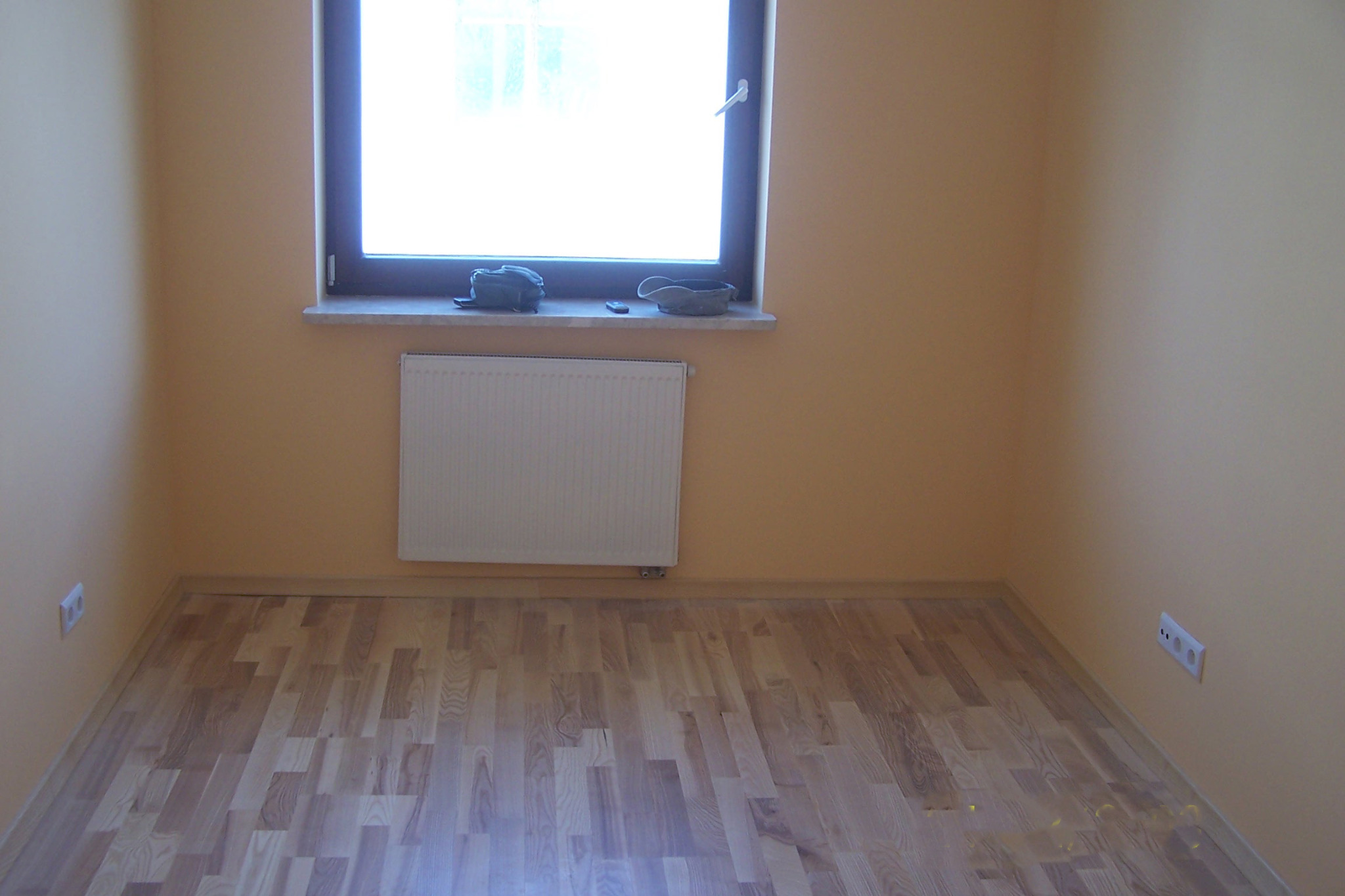 Pusta, pomalowana na beżowo ściana z oknem, grzejnikiem i drewnianą podłogą. Na parapecie dekoracje. Widoczne gniazdka elektryczne. Ujęcie z perspektywy podłogi.