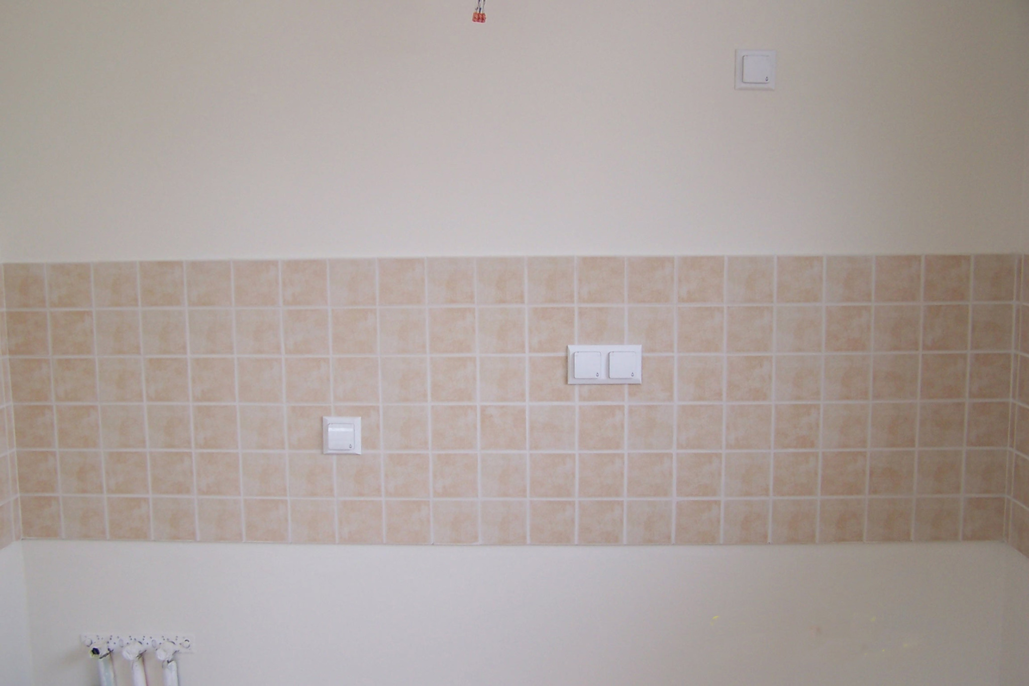 Beżowe płytki na ścianie z włącznikami światła. Wykończona ściana w łazience lub kuchni. Widoczne fugi i fragment grzejnika. Neutralne kolory.