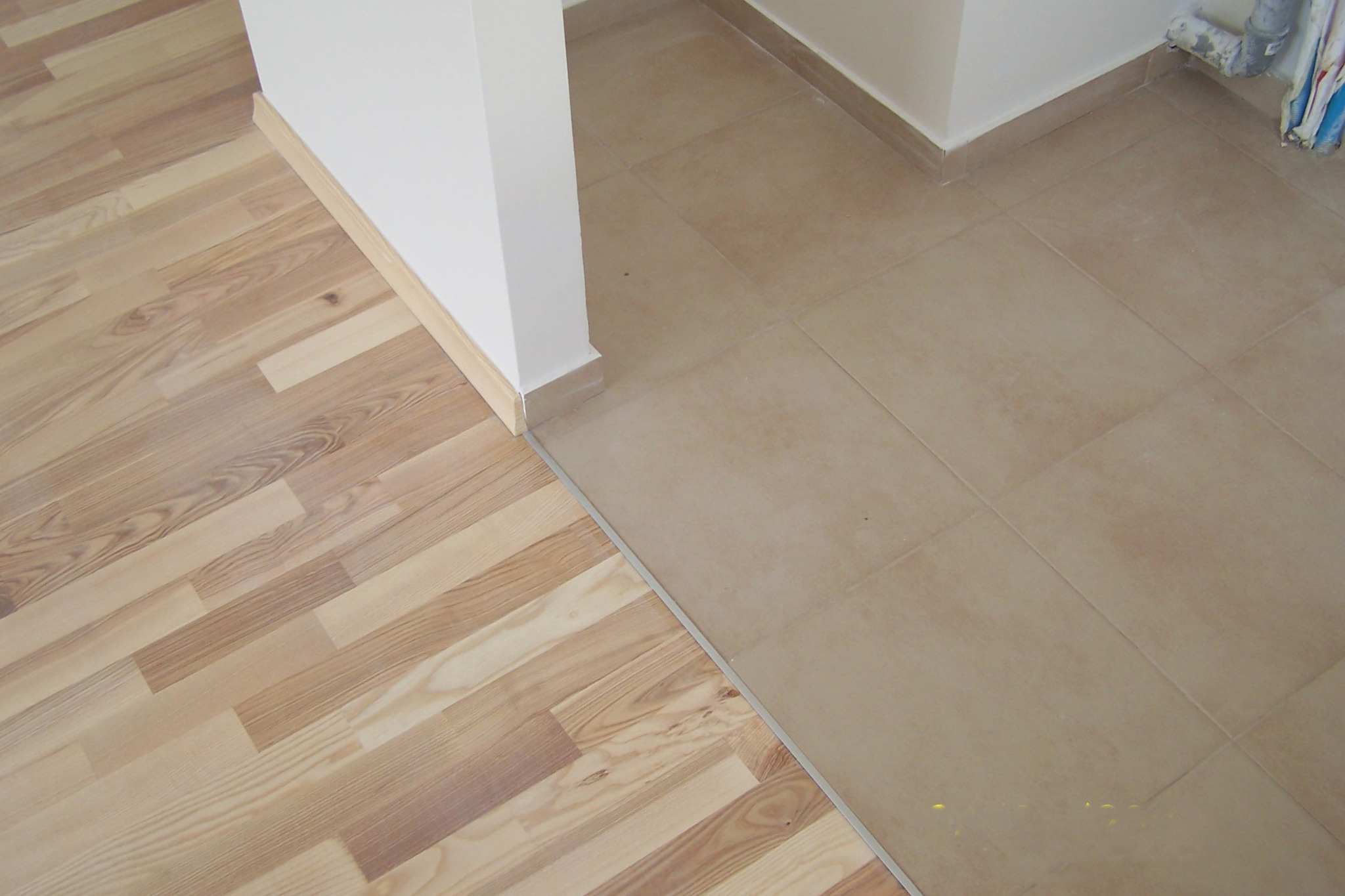 Połączenie parkietu i beżowych płytek podłogowych, oddzielonych listwą. Widoczne fragmenty białej ściany z listwami przypodłogowymi. Ujęcie z góry, naturalne światło.
