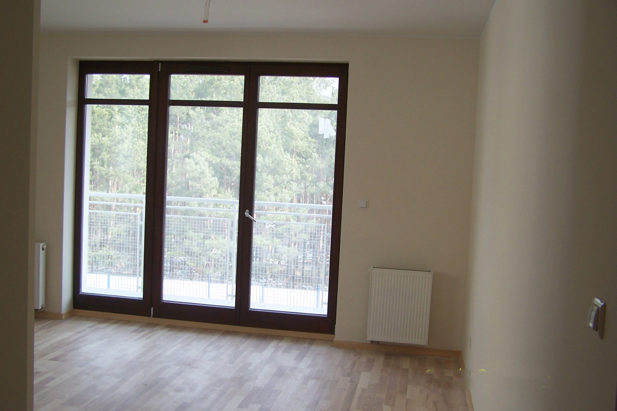 Jasne, puste wnętrze po remoncie z parkietem i dużym oknem balkonowym z widokiem na zieleń. Świeżo pomalowane ściany w odcieniach beżu. Grzejnik pod oknem.