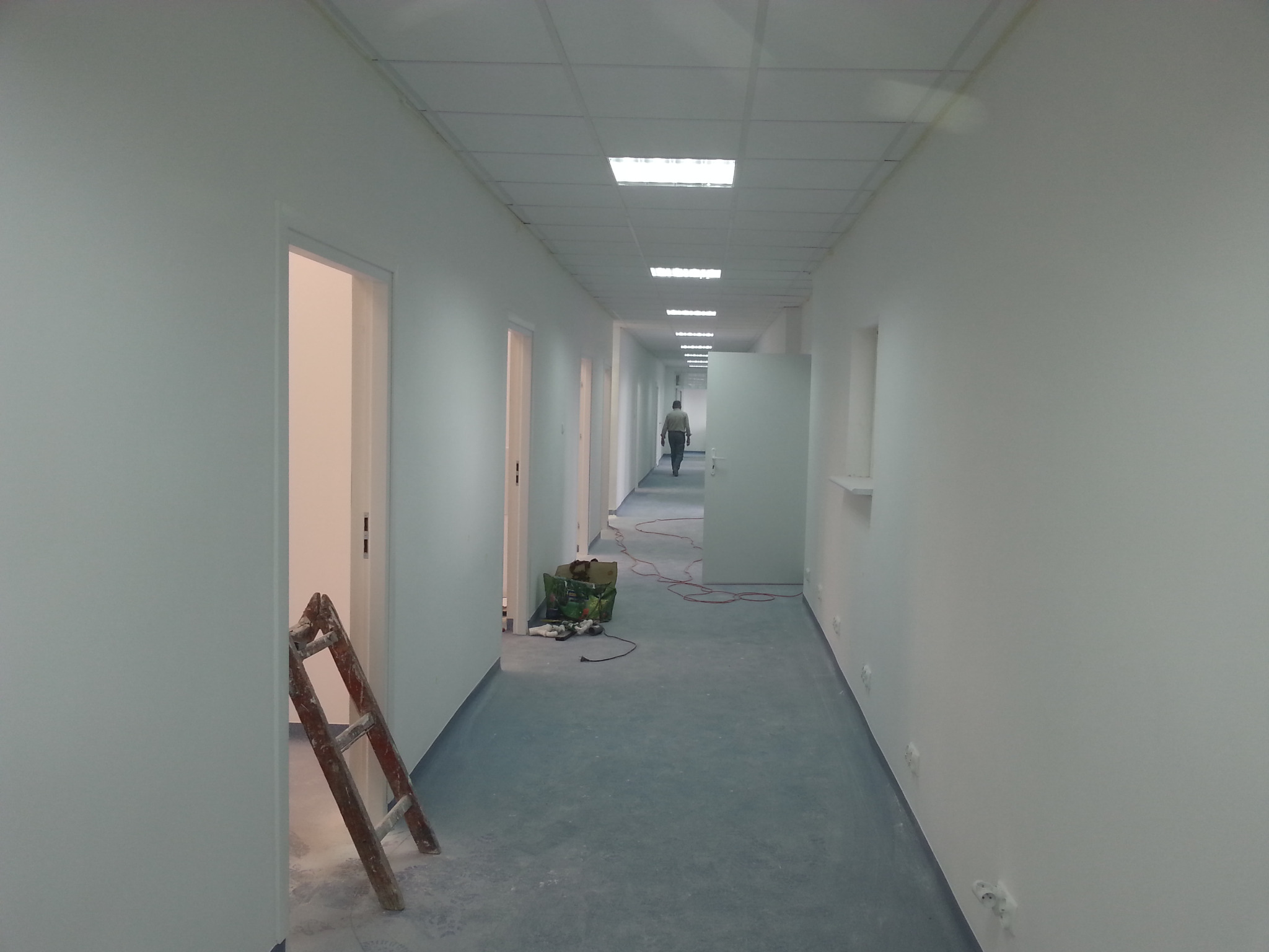 Biały korytarz biurowy w trakcie prac wykończeniowych. Widoczna drabina, narzędzia i osoba w oddali. Oświetlenie sufitowe i świeżo pomalowane ściany.