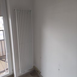 Ark - Biały grzejnik panelowy zamontowany przy oknie balkonowym na tle białej ściany z widocznymi śladami po instalacji. Jasne panele podłogowe.