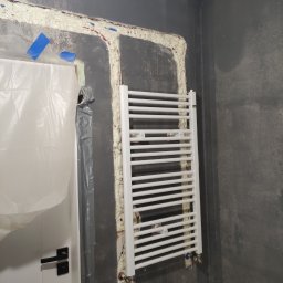 Ark - Biały grzejnik drabinkowy zamontowany na szarej ścianie w trakcie prac wykończeniowych. Widoczne pianka montażowa, narzędzia i zabezpieczona folią biała ościeżnica drzwi.