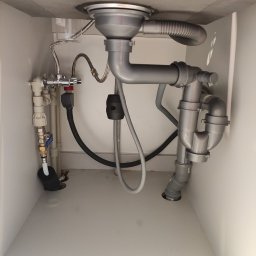 Ark - Szare rury odpływowe i zawory pod zlewem w białej szafce. Widoczne złącza, wężyki i syfon. Ujęcie z dołu, ukazujące instalację hydrauliczną.