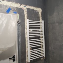 Ark - Biały grzejnik łazienkowy zamontowany na szarej ścianie, z widoczną pianką montażową wokół rur. W tle zakryte folią drzwi i wiertarka na podłodze.